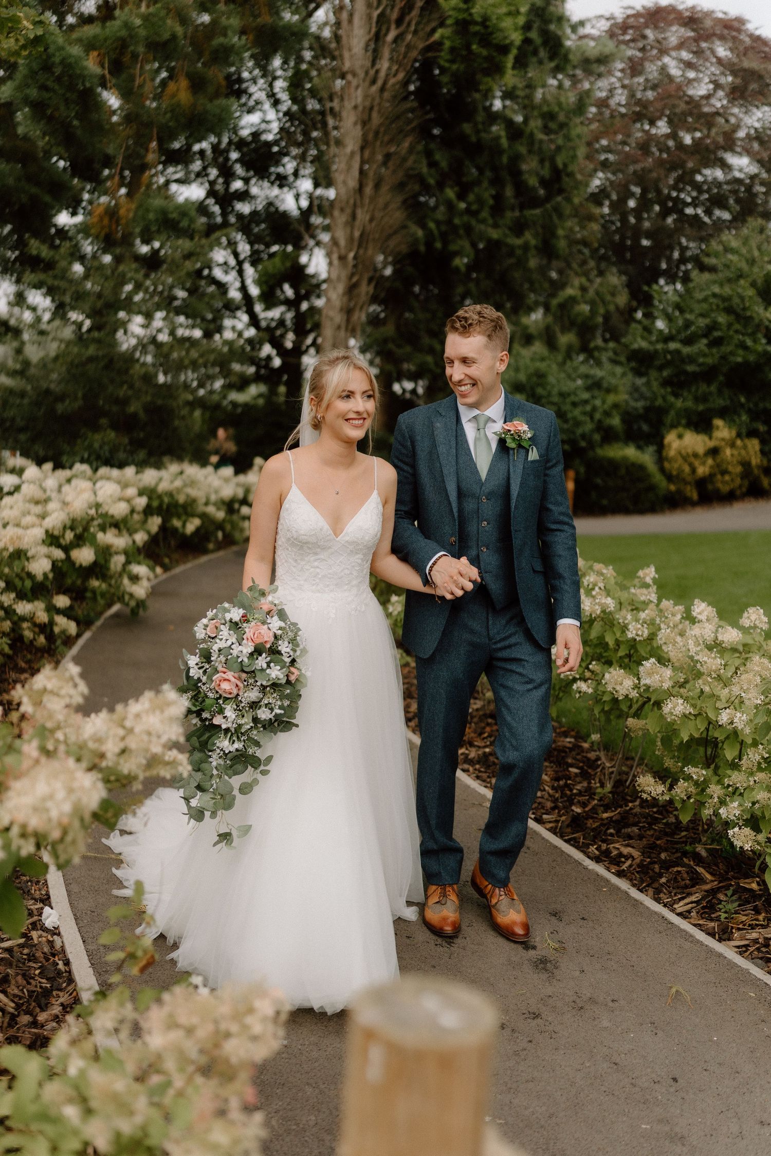 A couple walks along a garden path on their wedding day, with the bride in white and groom in navy blue at Hothorpe Hall.