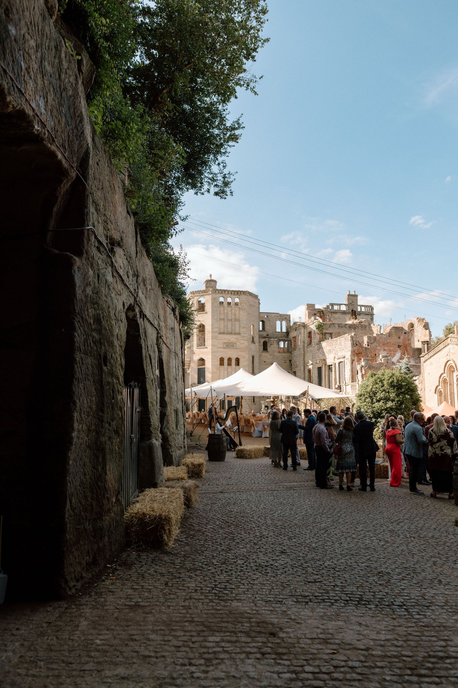 A cobblestone street lined with castle-like buildings hosts an outdoor event with white tents and mingling guests at Guys Cliffe in Warwick.