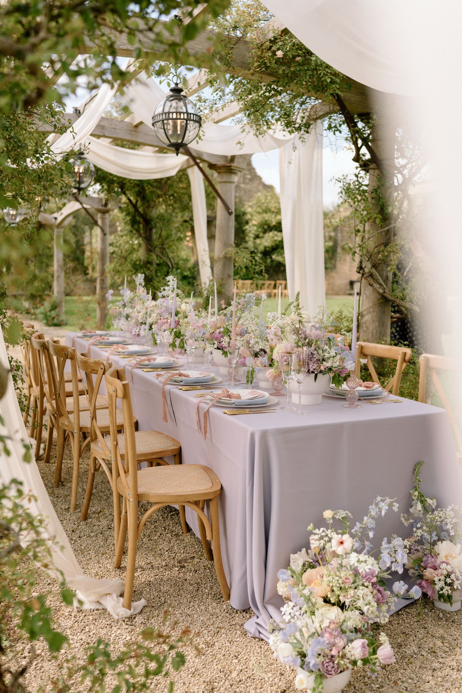 A dreamy outdoor wedding reception table with white draping and wooden chairs under hanging lanterns.