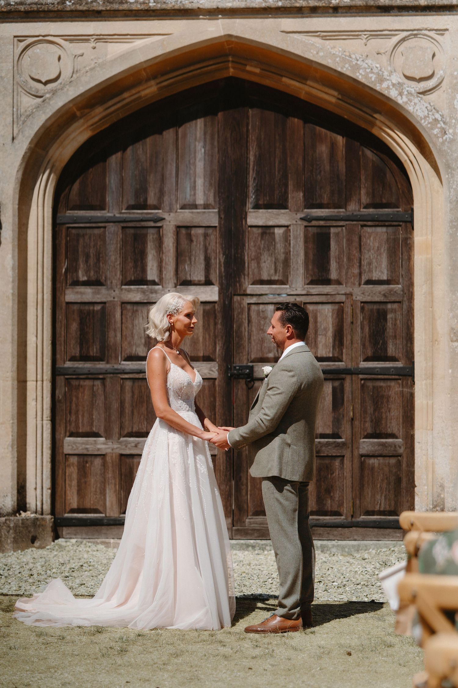 Bride and groom hold hands smiling before grand wooden arched doors at a historic stone venue wedding.