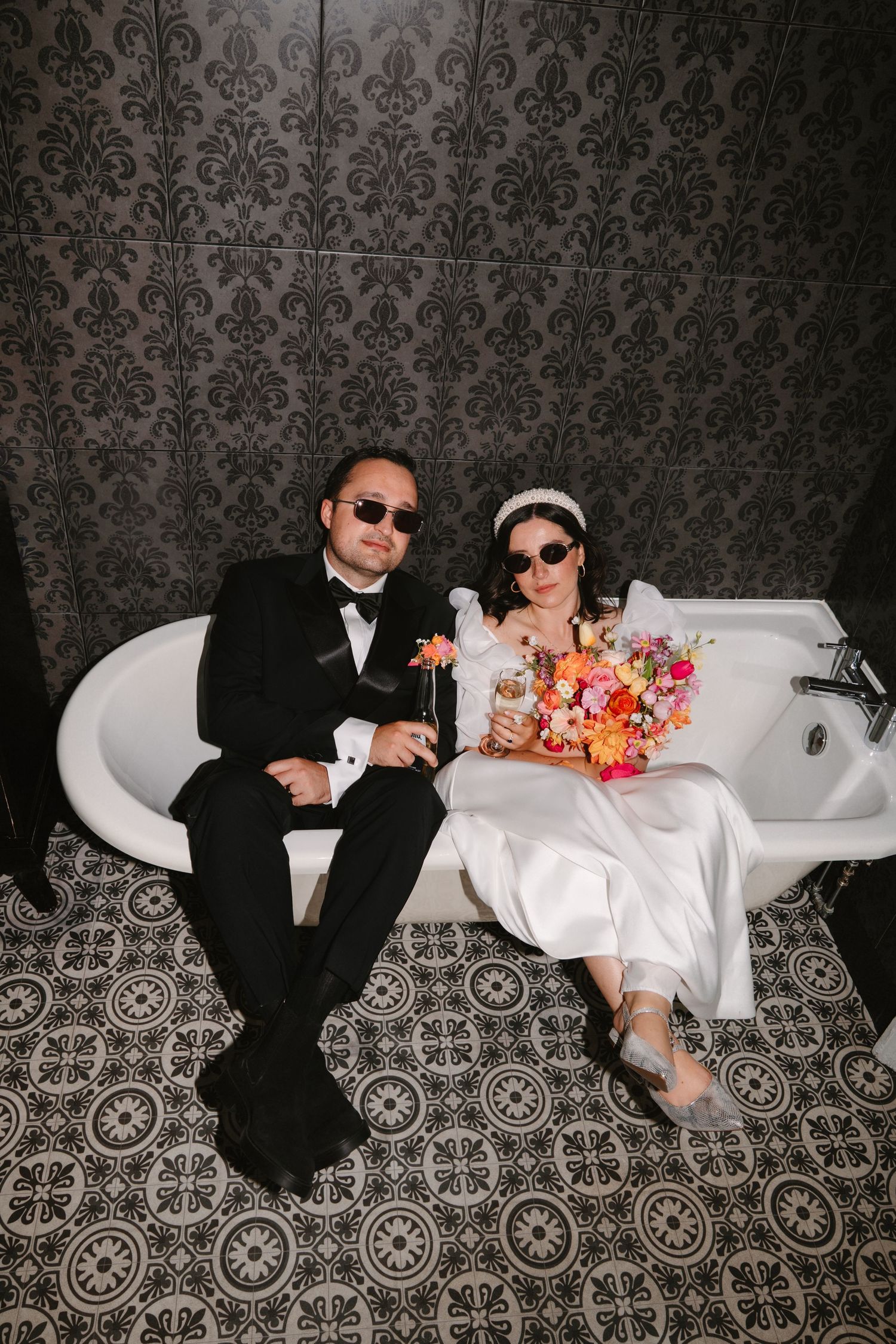 Bride and groom in sunglasses sitting in a vintage bathtub, holding drinks and colorful bouquet in a dark patterned bathroom.