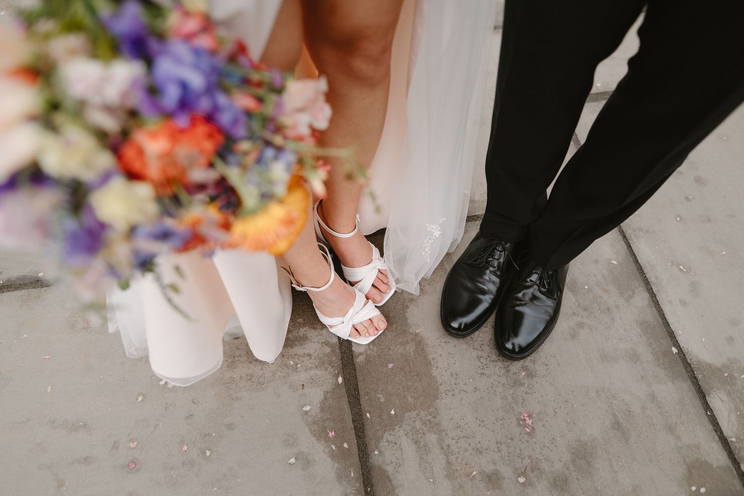 Close-up of bride in white heeled sandals and groom in black dress shoes standing together with colorful wedding bouquet.