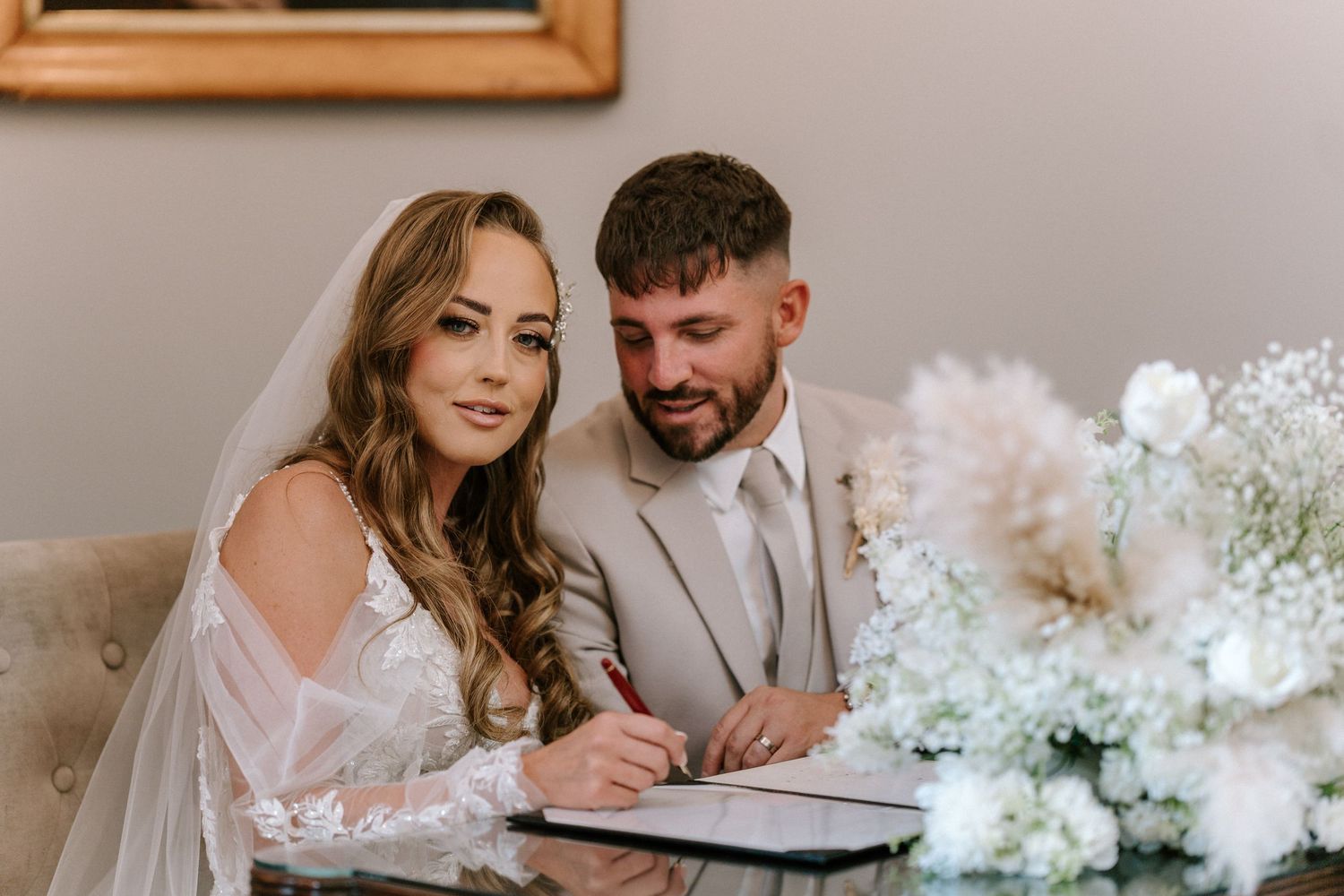 A couple at their wedding celebration writing in a guestbook surrounded by white floral decorations.