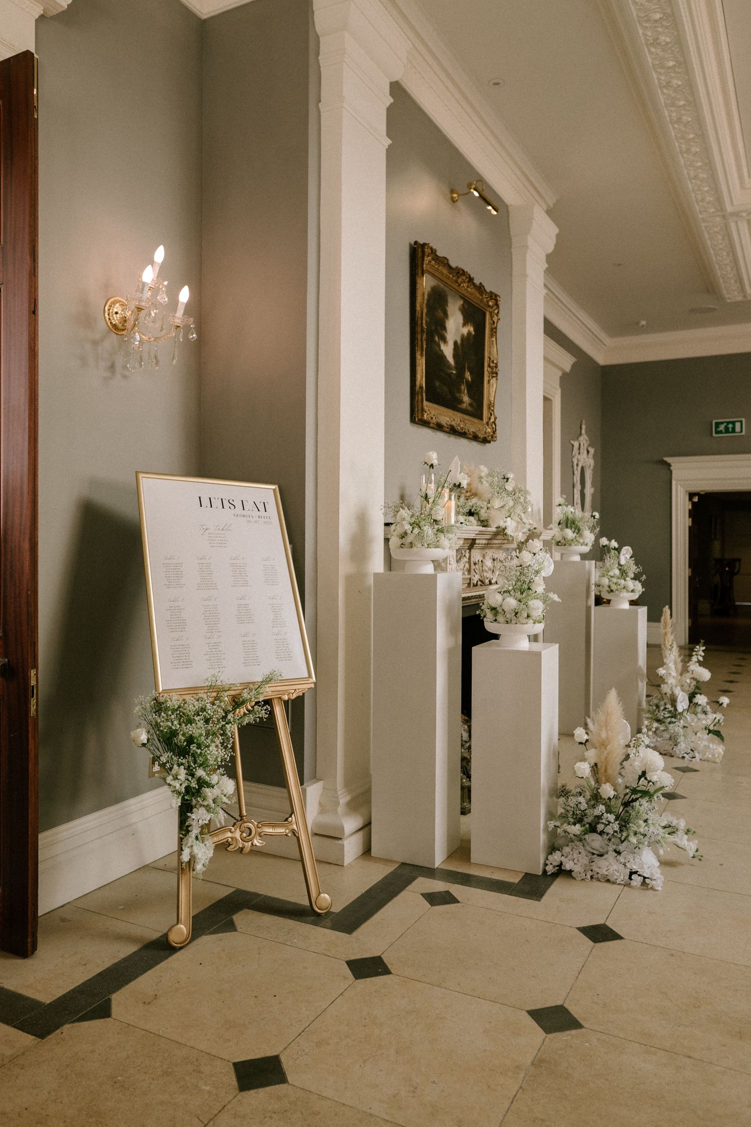 Elegant hallway decorated with white floral arrangements, candles, and display boards for a wedding reception.
