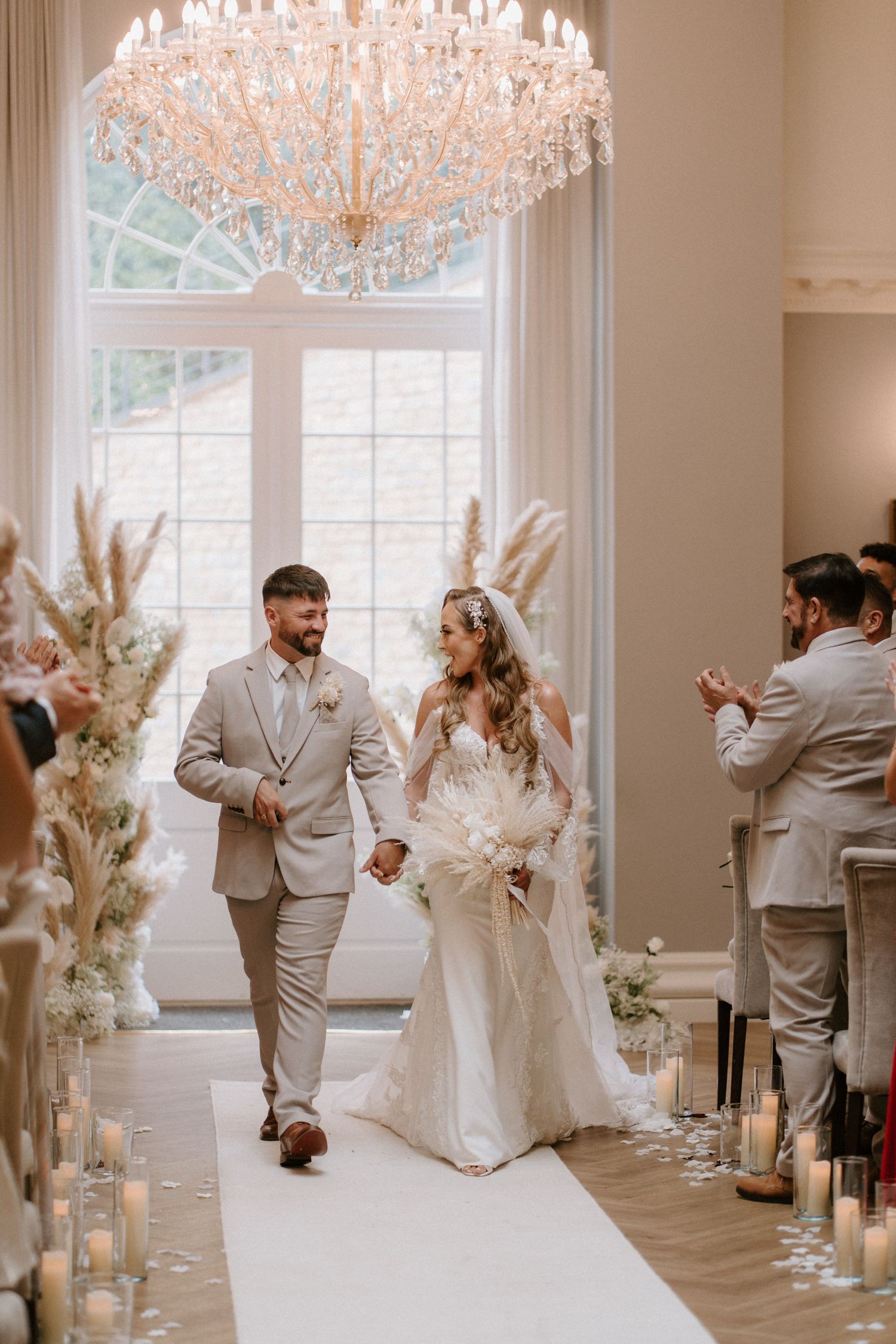 Wedding ceremony in elegant room with crystal chandelier and candle-lined aisle.