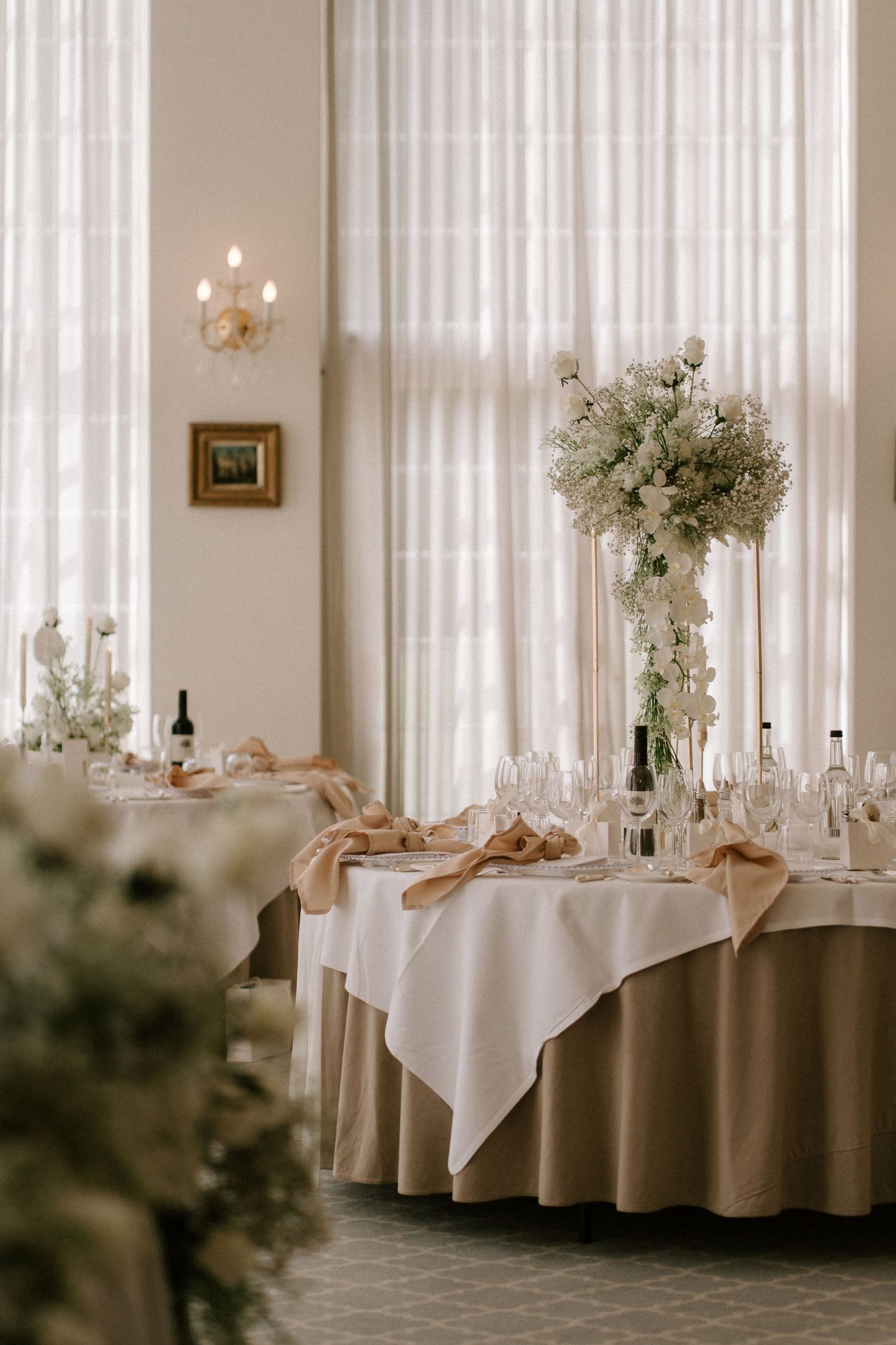 Elegant reception table setting with tall white floral centerpiece, white linens, and sheer curtained windows.