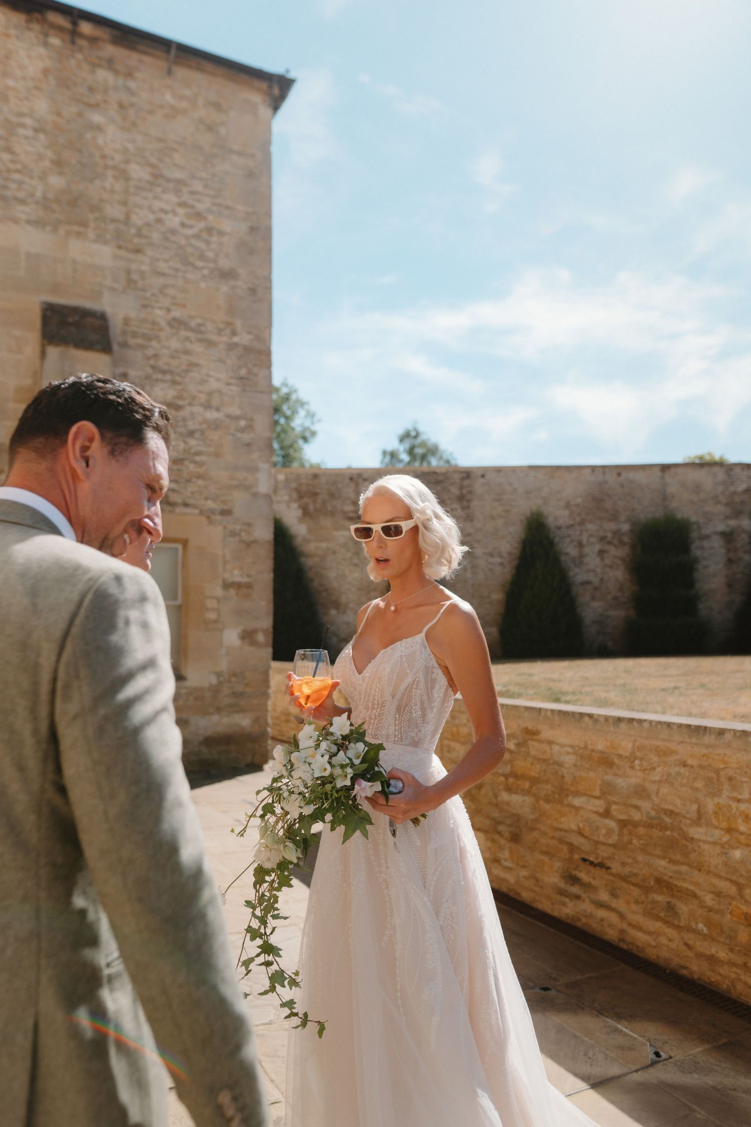 Bride in white gown and sunglasses holds bouquet and champagne beside groom in gray suit at sunny stone venue.