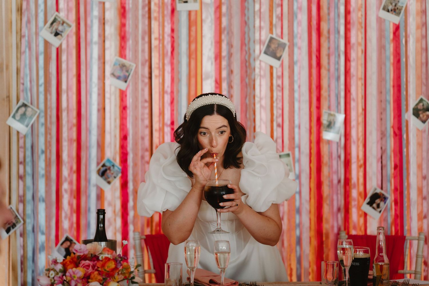 Bride in white dress sipping drink at colorful streamer backdrop with floating polaroids at festive wedding reception.