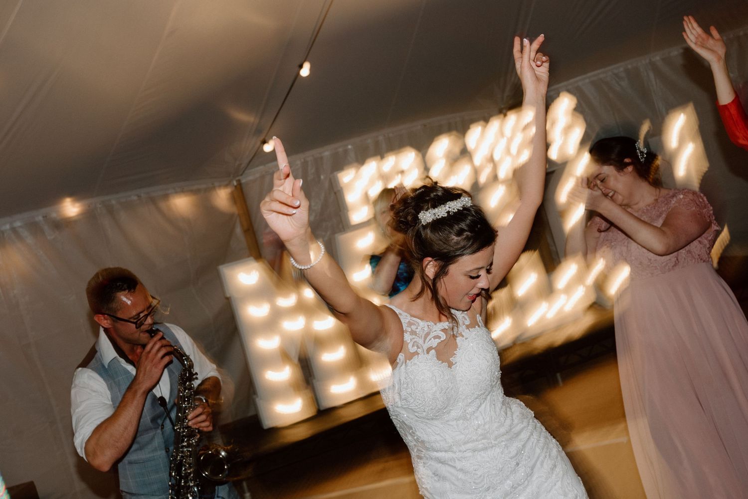 A wedding celebration on the dance floor with glowing lights in the background and dancing with raised arms.