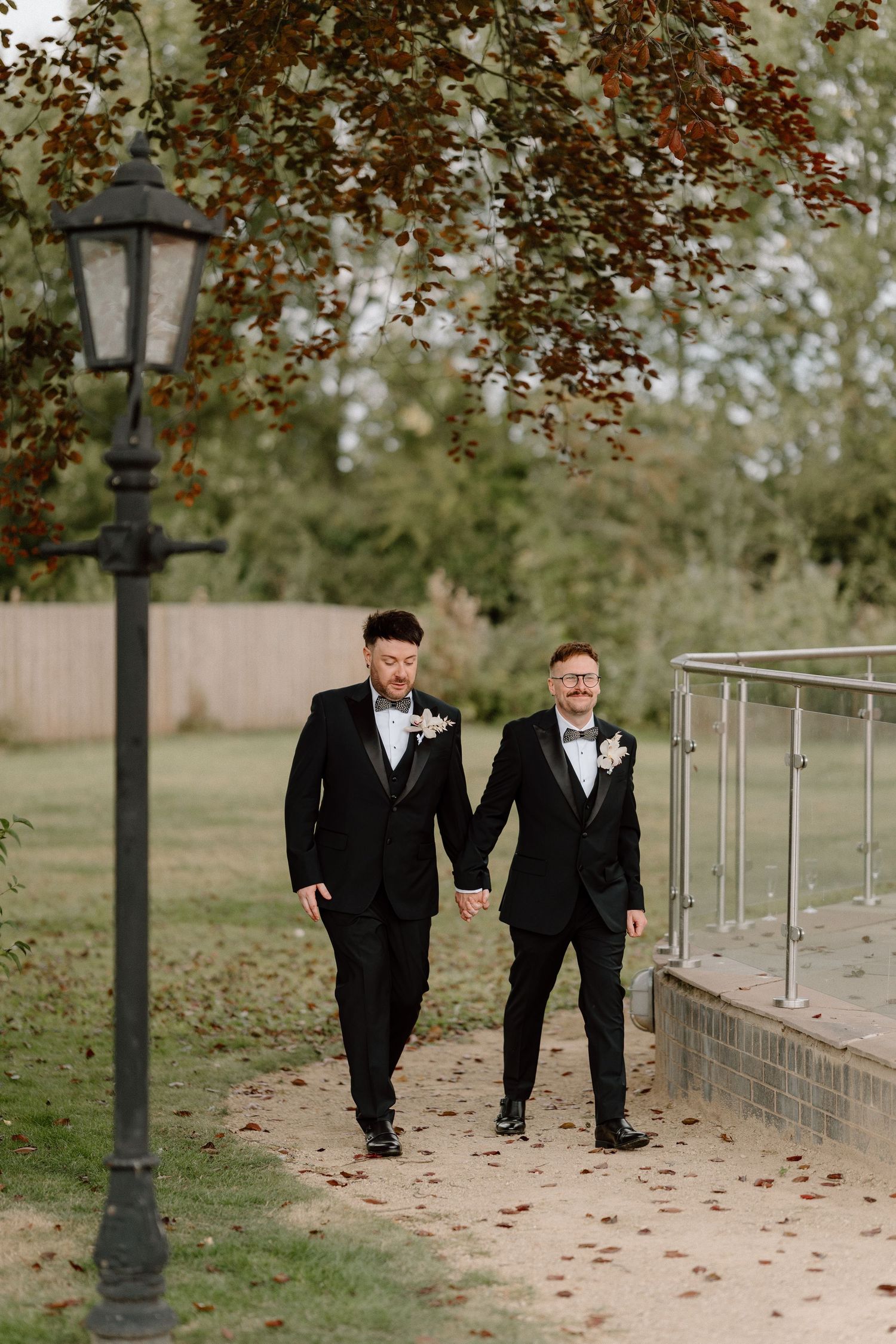 Two people in black tuxedos walking hand in hand along an autumn path with vintage lamppost at The Manor House at Quorn.
