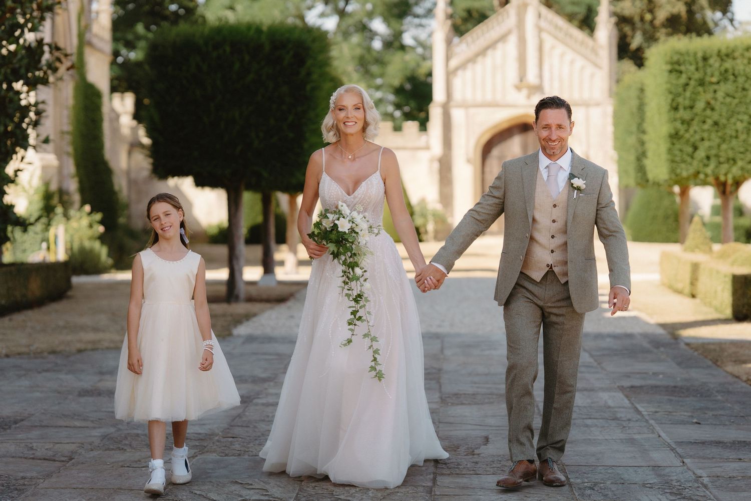 Bride and groom walking with flower girl at elegant outdoor wedding venue with manicured hedges.