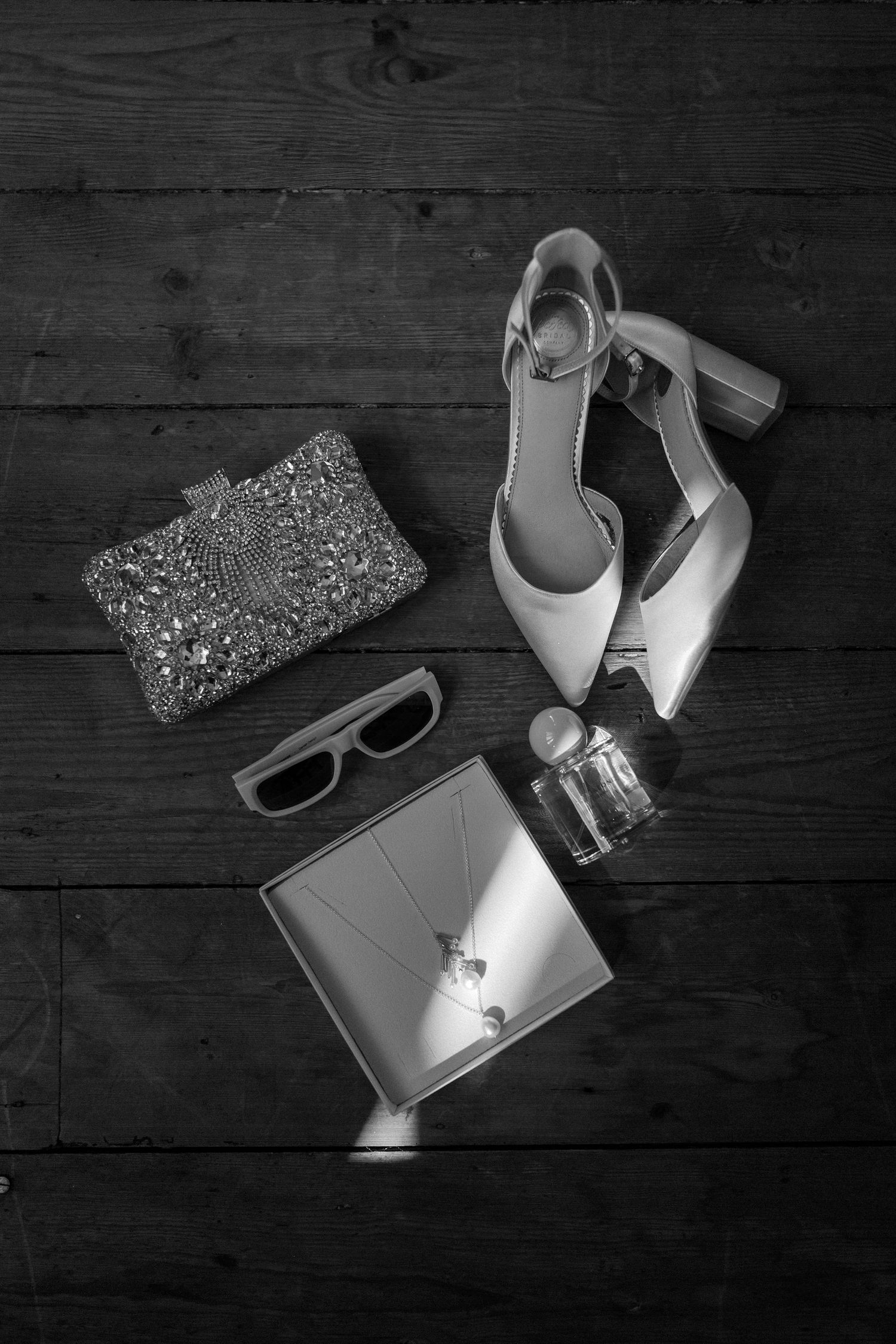 Black and white flat lay of elegant heels, embellished clutch, sunglasses, perfume, and jewelry box on wood floor.
