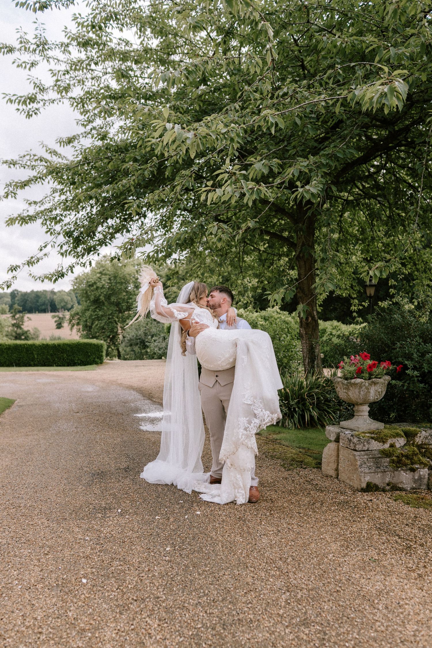 Wedding celebration group dances down tree-lined gravel path on sunny day.