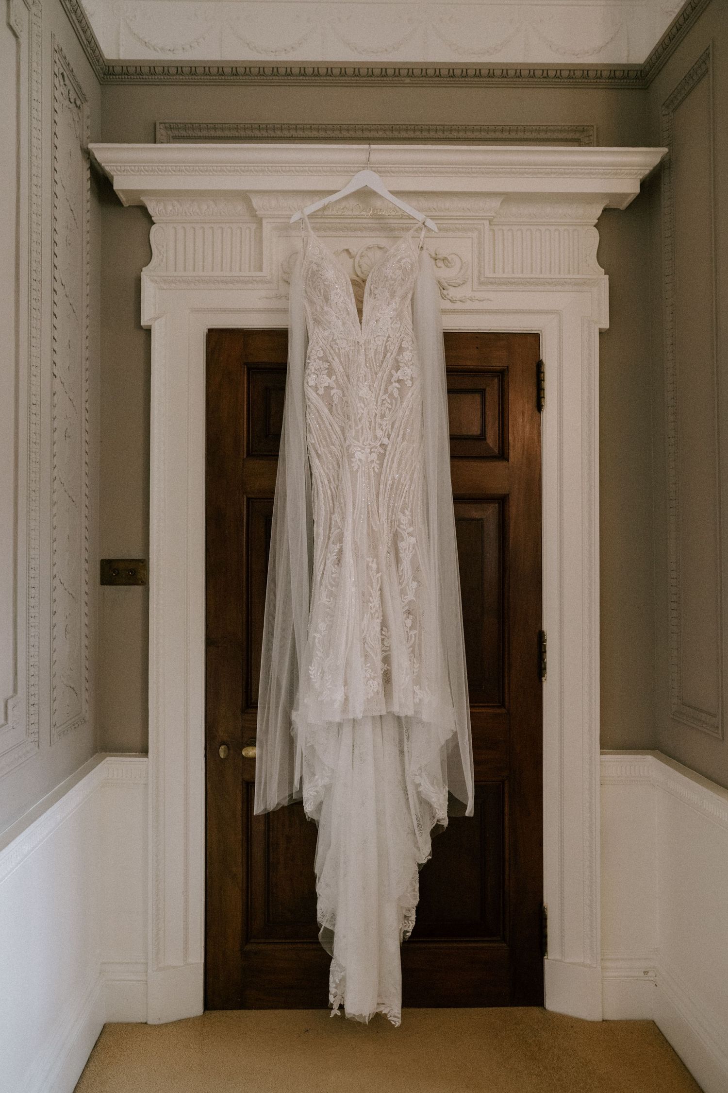 An elegant white lace wedding dress with intricate details hangs on display against wooden double doors.