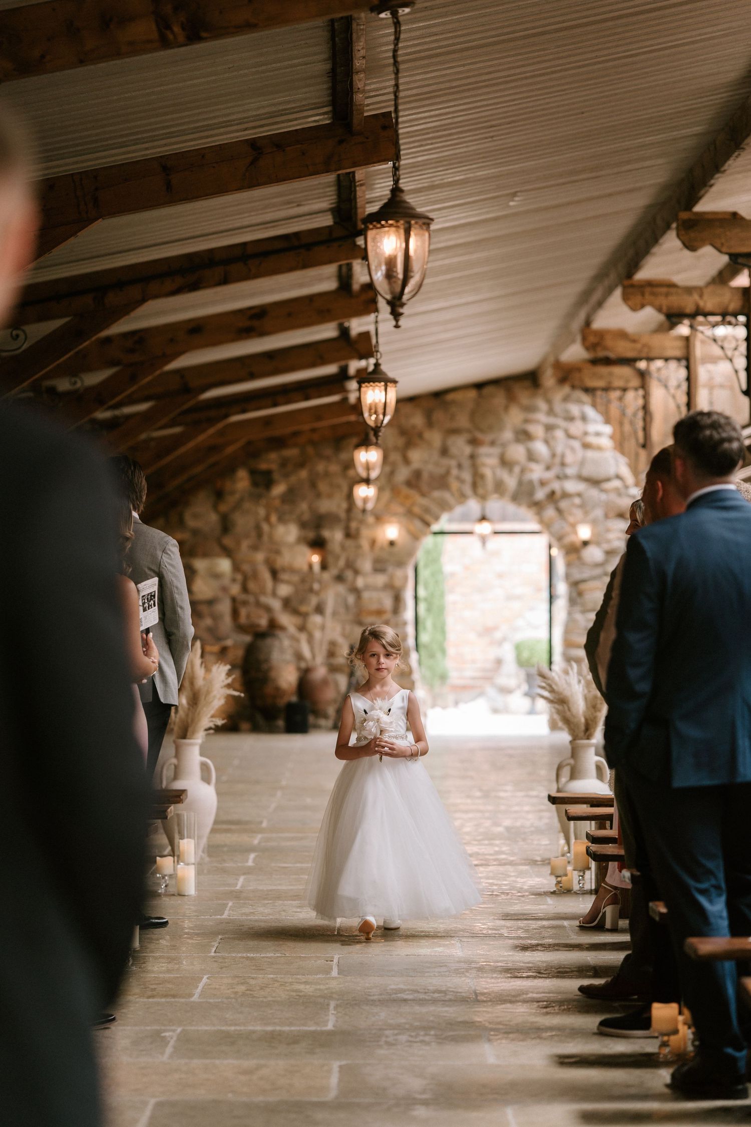 A stone hallway with hanging lanterns during a wedding ceremony with guests seated along the sides.