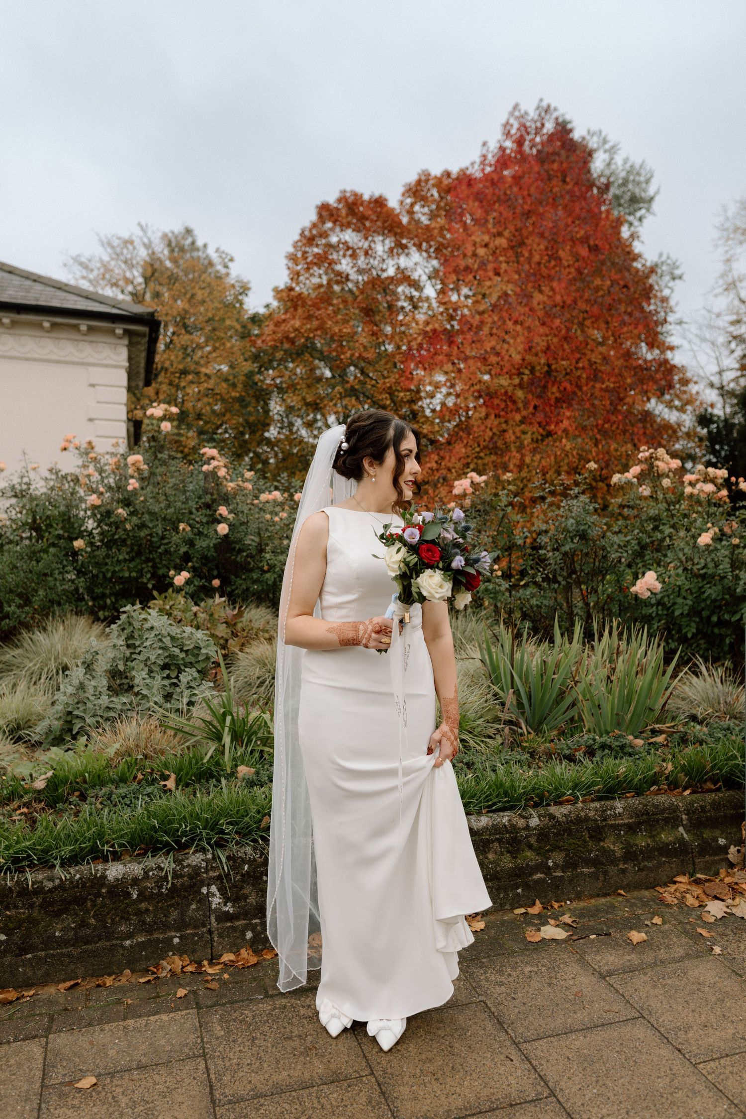 A bride in white stands in a garden with autumn foliage holding a bouquet at The Glasshouse in Leamington Spa.