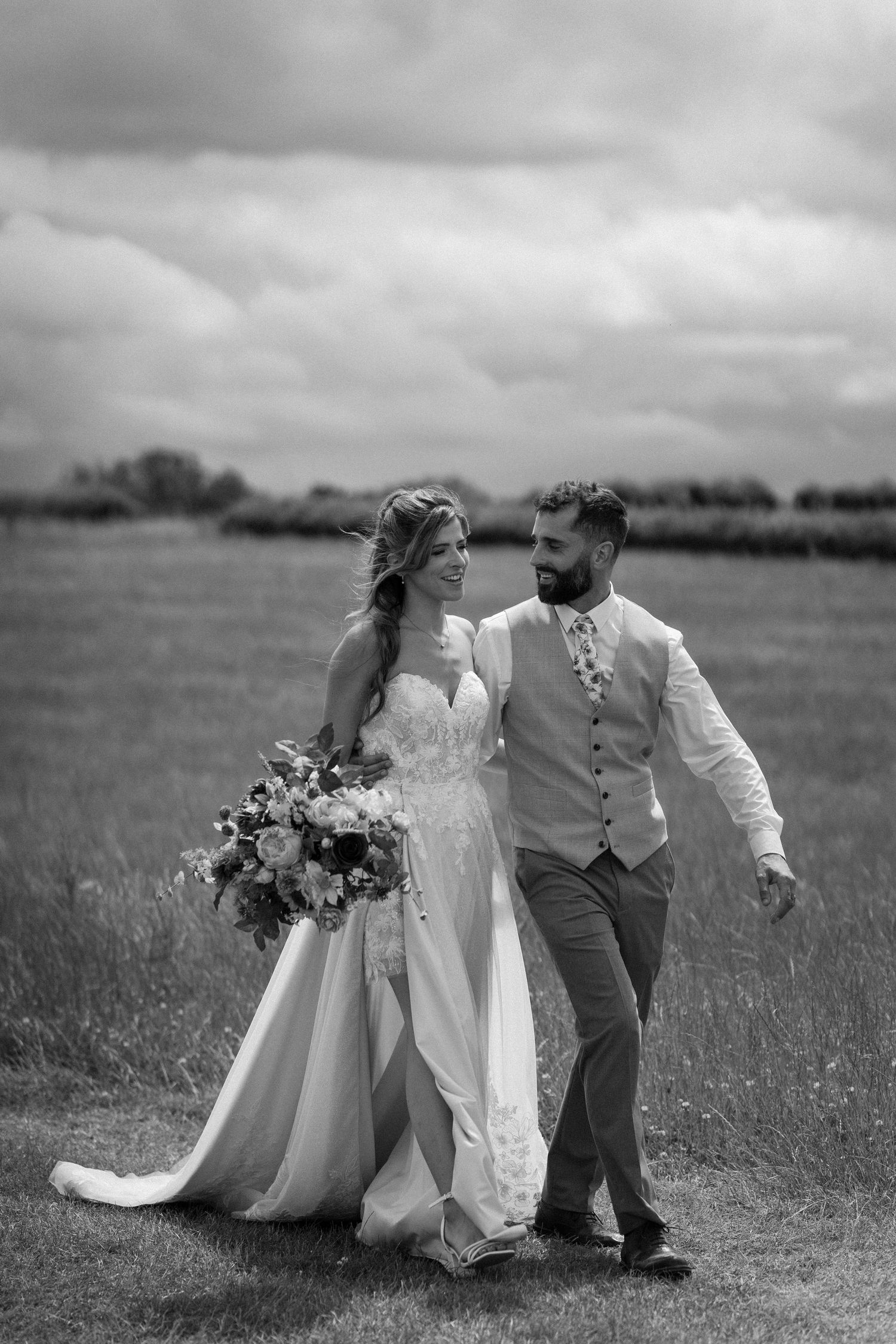 Black and white photo of a bride and groom walking through a field, smiling at each other on their wedding day.
