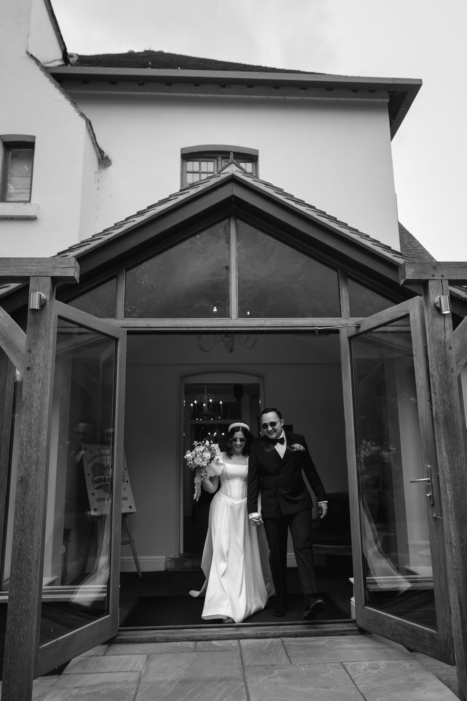 Black and white photo of a bride and groom exiting a glass-doored venue on their wedding day.
