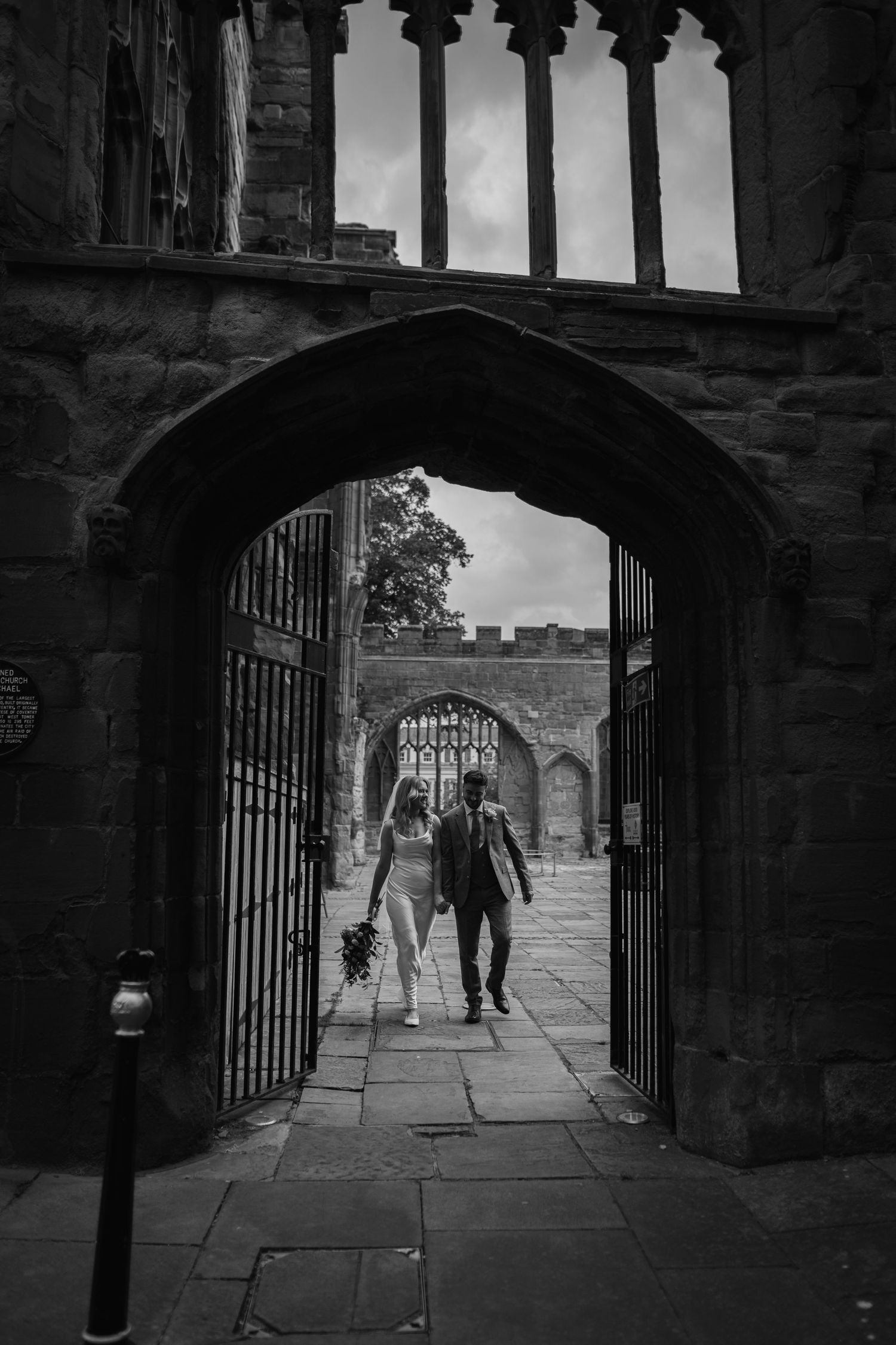 A couple walking through a gothic stone archway with their dog in black and white at Coventry Cathedral.