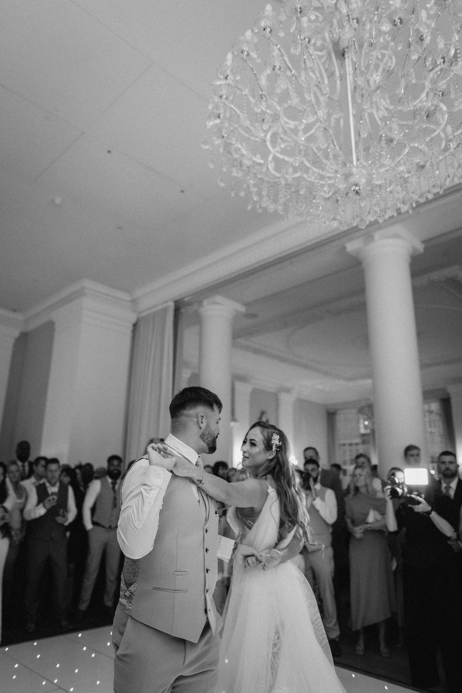A couple shares their first dance under a crystal chandelier while guests watch in a grand ballroom.