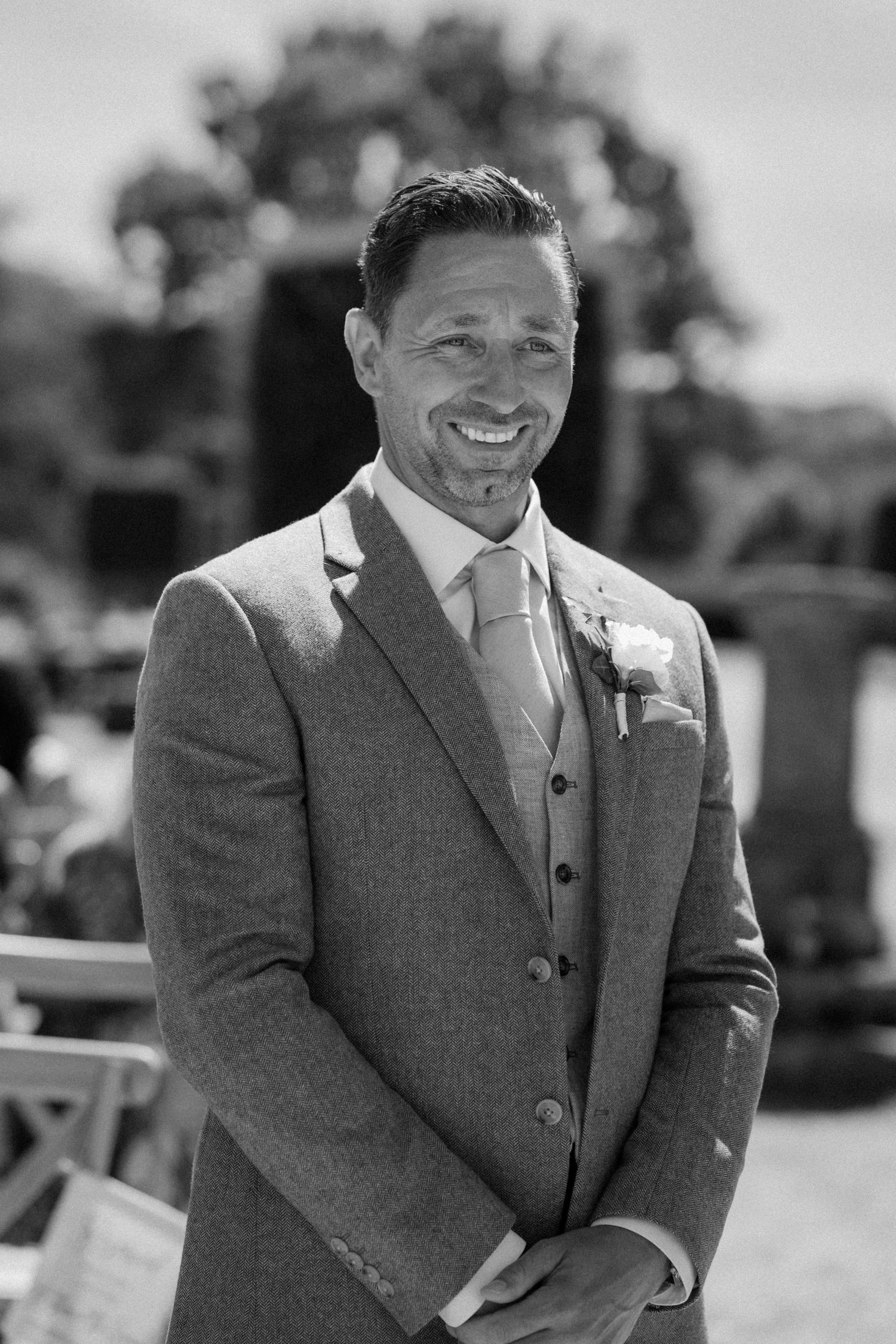 Smiling groom in elegant grey tweed suit with boutonniere, standing outdoors at wedding ceremony.