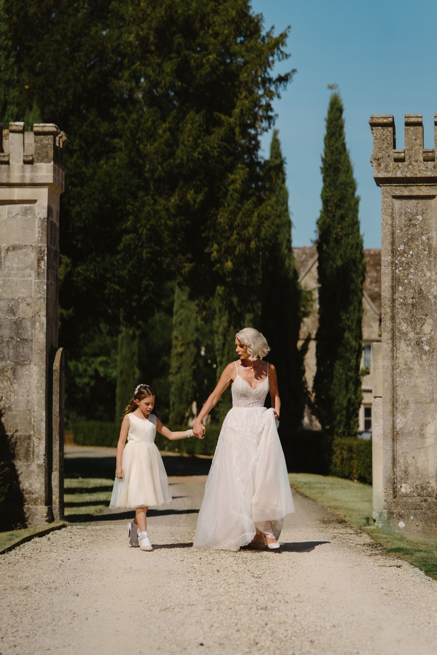 Bride in a flowing white gown holds hands with a flower girl walking through stone gates lined with cypress trees.