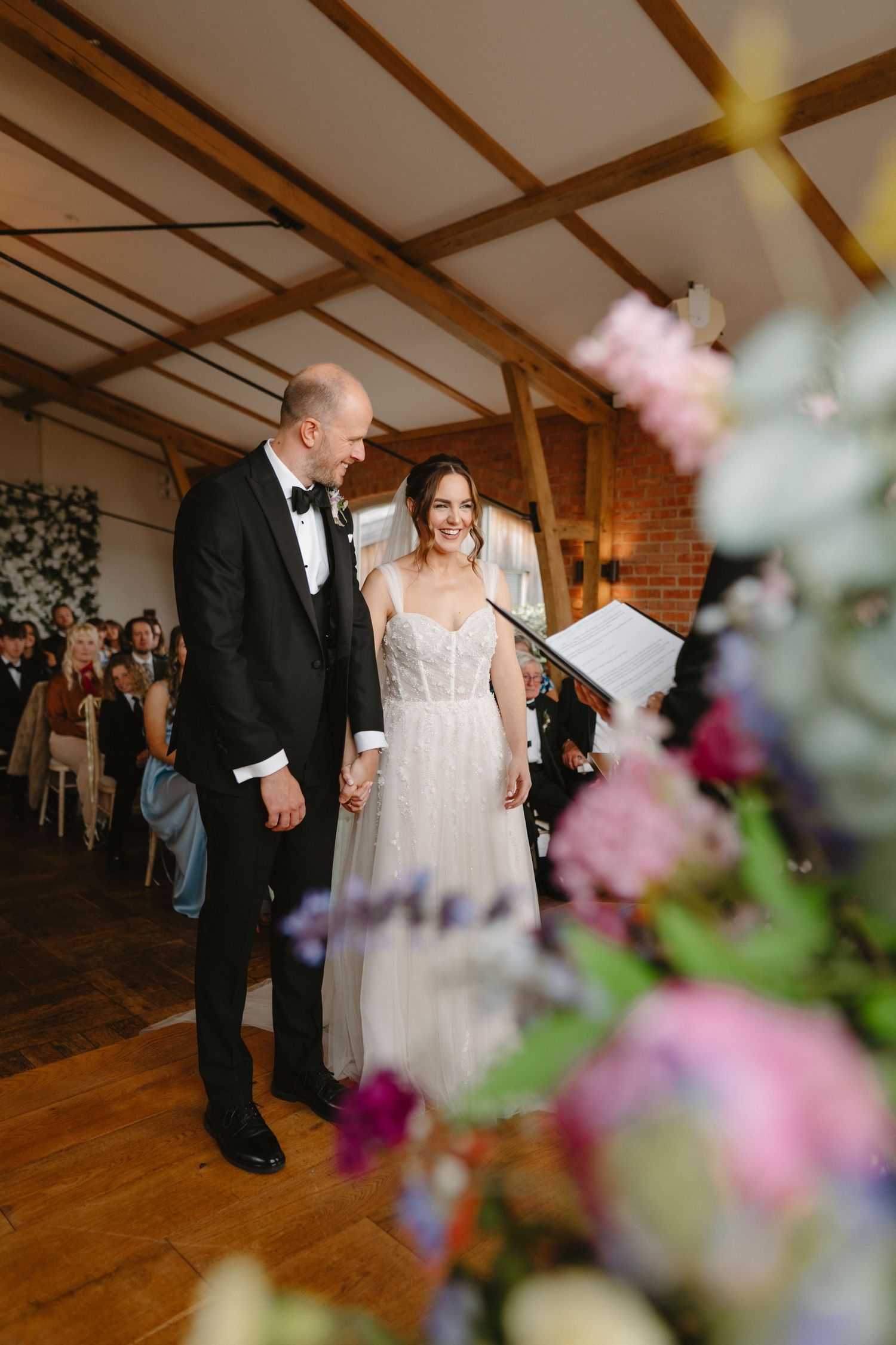 Bride and groom smiling together during wedding ceremony in rustic barn venue with exposed beams and floral arrangements.