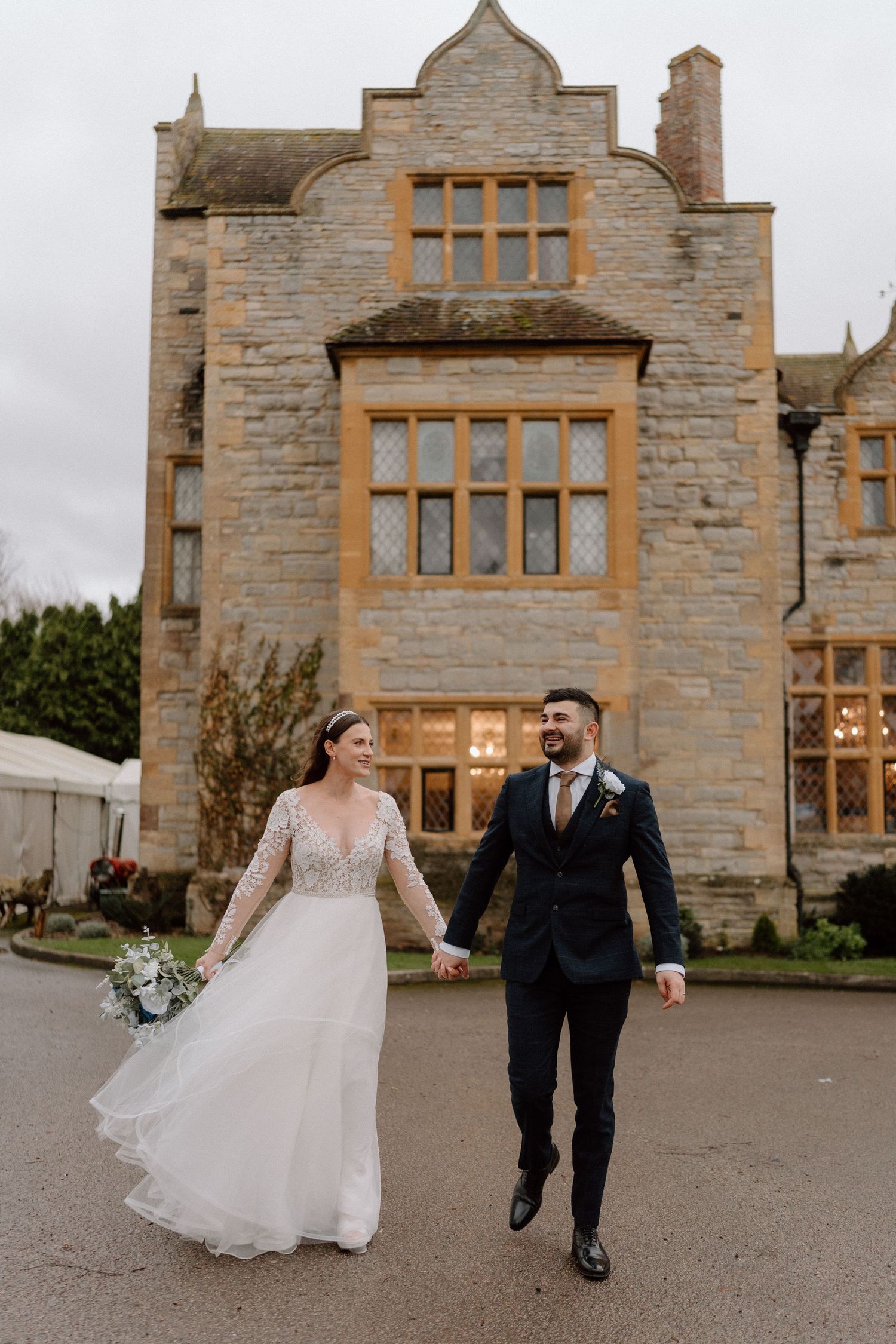 A couple walks hand in hand outside a grand stone manor house.