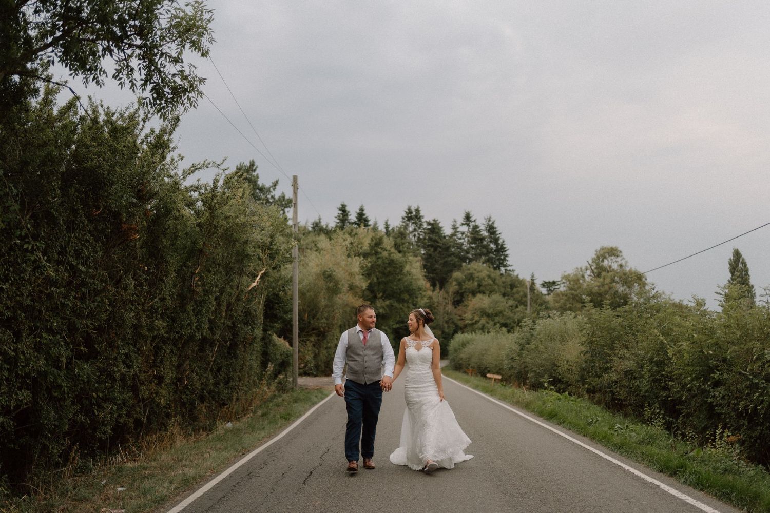 Two people holding hands walk together down empty rural road on cloudy day with trees lining both sides.