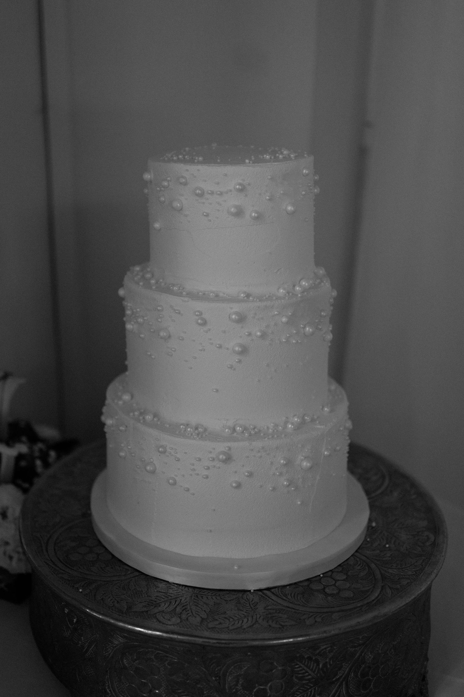 A classic three-tiered white wedding cake decorated with delicate pearl-like dots sits elegantly on a silver cake stand at Warwick House.