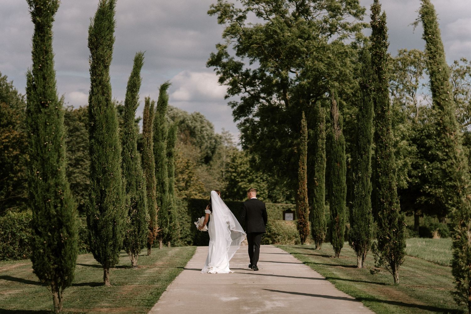A married couple walking down a cypress tree-lined pathway under cloudy skies.