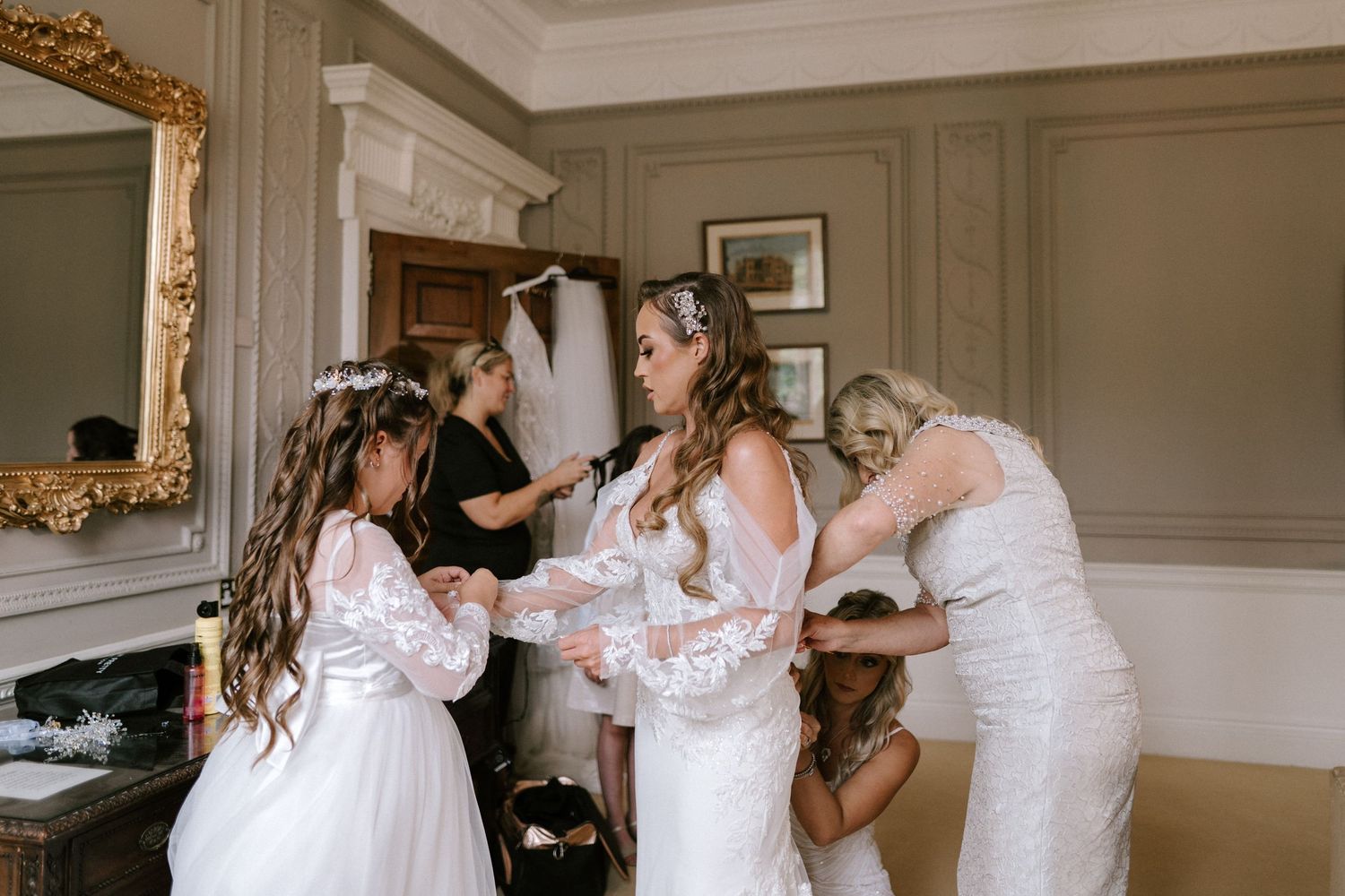 A bride getting dressed and prepared with help from others in an elegant room with ornate gold mirror.