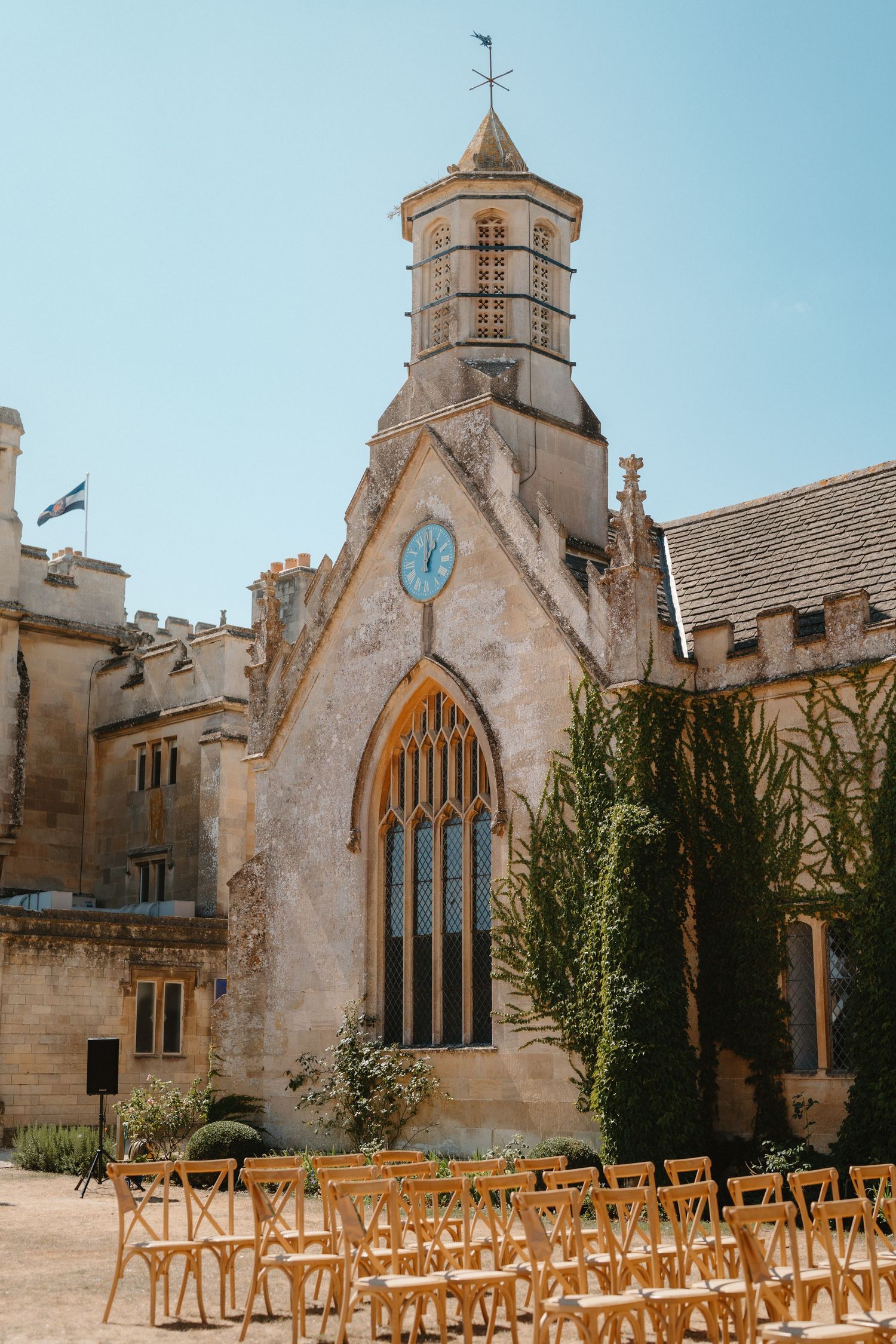 Outdoor wedding ceremony chairs arranged before a historic stone chapel with clock tower under clear blue sky.