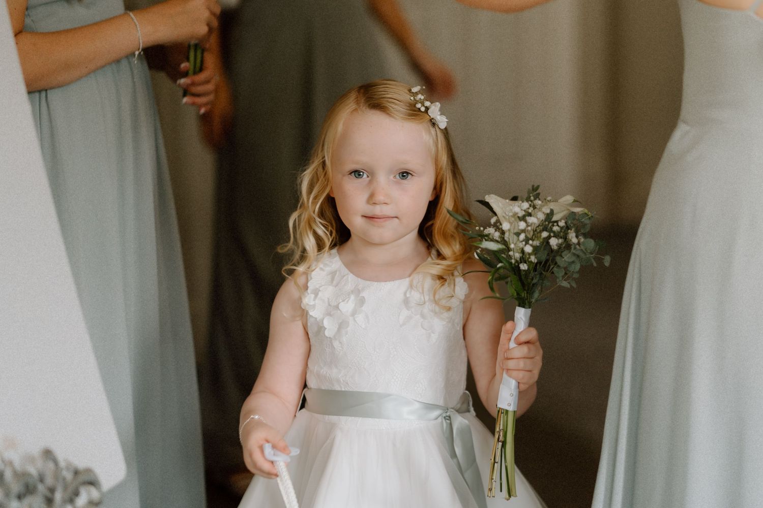 Young flower girl in white dress with silver sash stands among curtains and holds delicate white flowers at wedding at Warwick House.