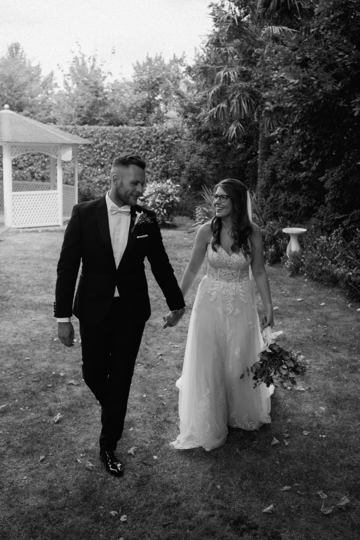 A black and white wedding photo showing a couple walking together in a garden with a gazebo visible in the background at Warwick House.