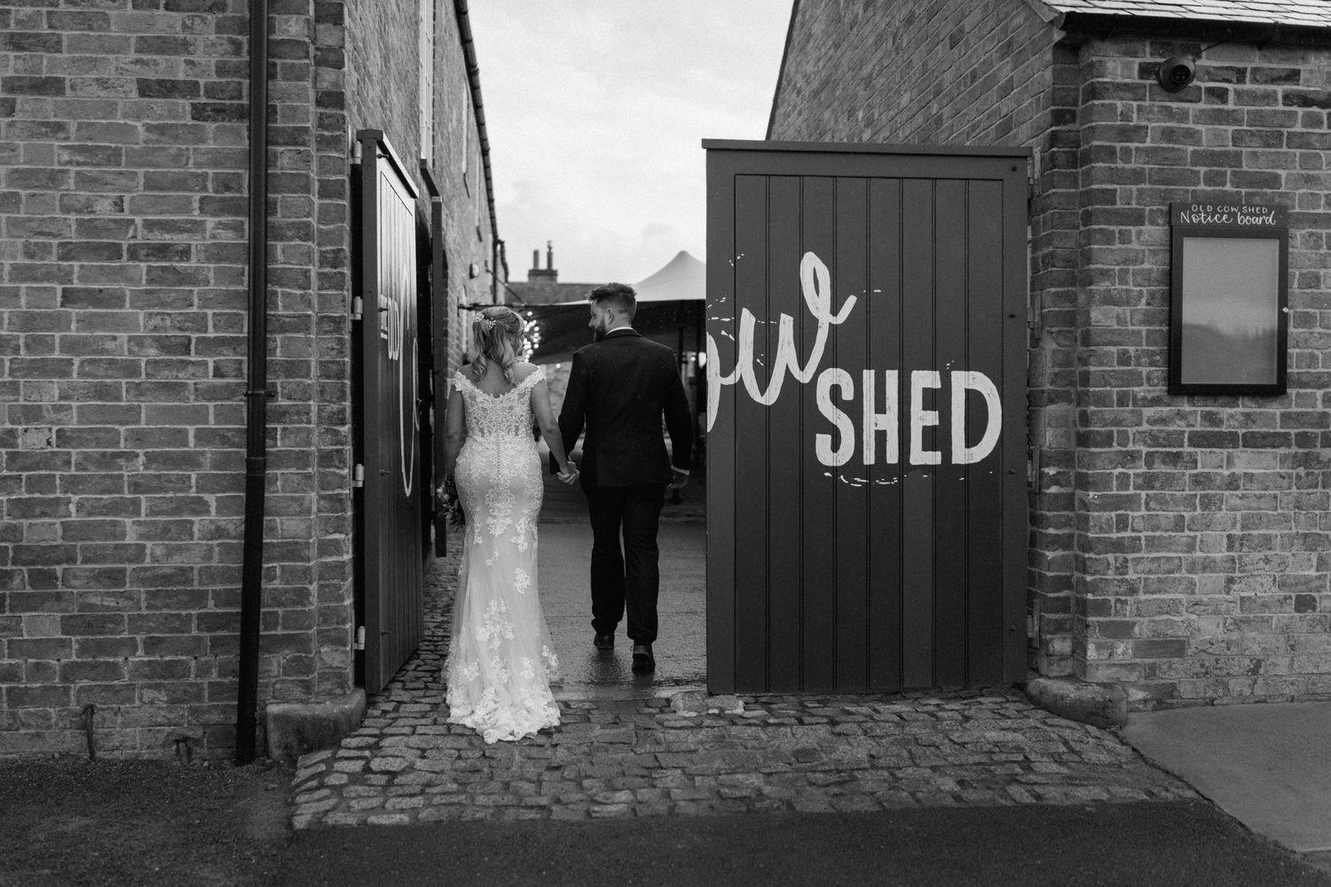 Black and white wedding photo of couple standing by rustic door labeled 'The Shed' at The Old Cow Shed.
