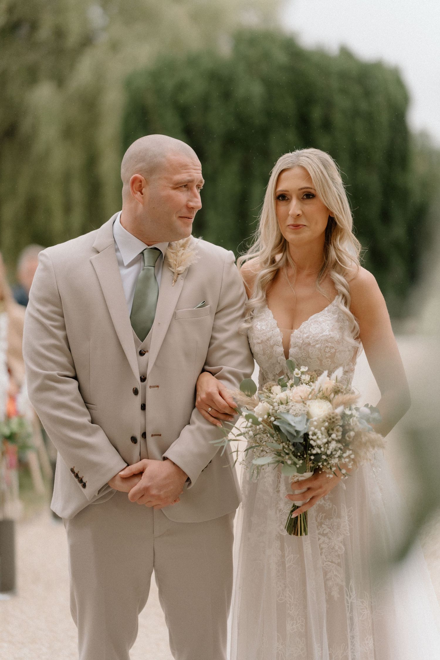 Couple in neutral toned wedding attire posing together outdoors with willow trees in background in Warwickshire.