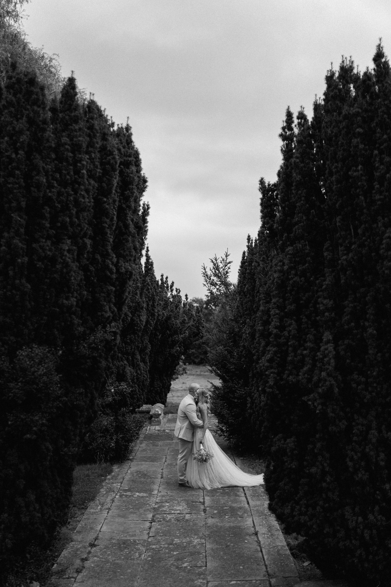 Black and white aerial view of couple walking between tall cypress trees on stone path in Warwickshire.