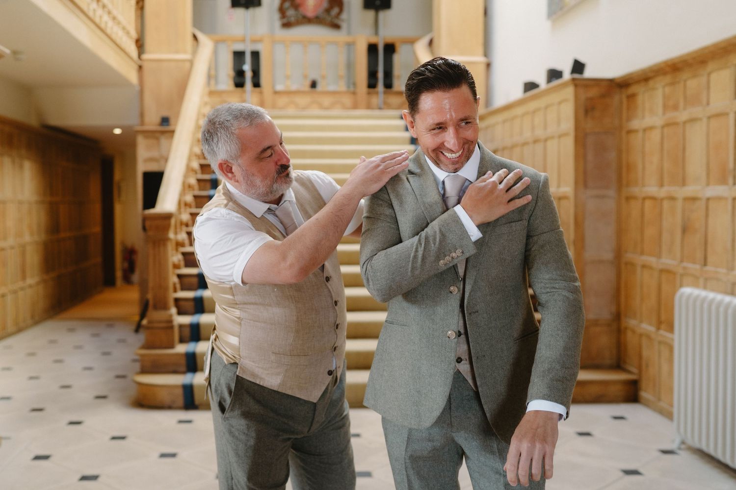 Two men laughing together in an elegant wood-paneled hall, one adjusting the other's suit jacket collar.