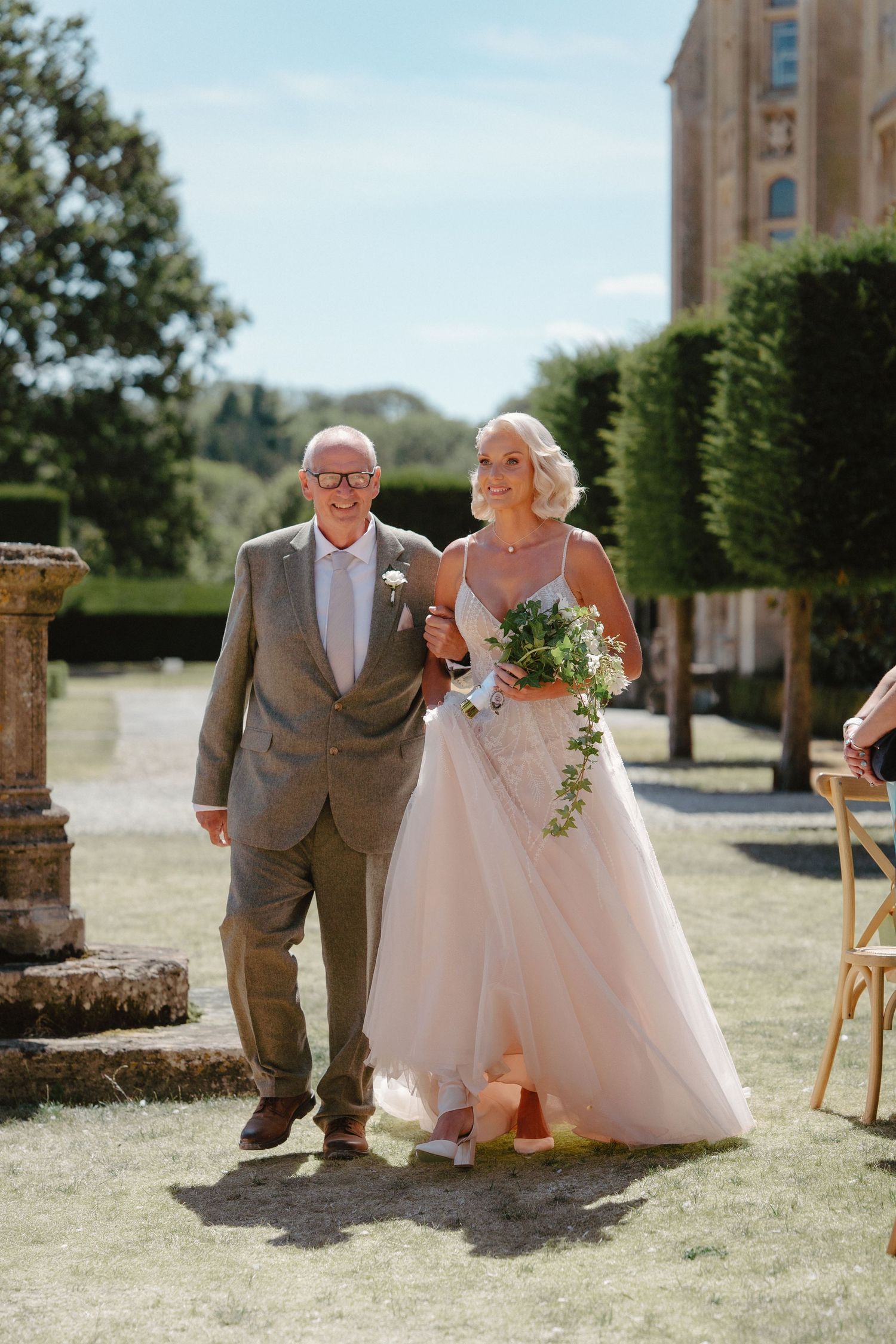 Bride in blush tulle gown walks down outdoor aisle with older gentleman in grey suit at English estate wedding.
