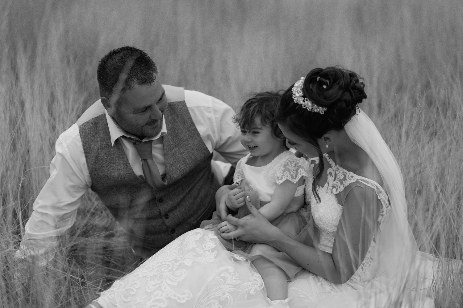 A black and white wedding photo shows a family sitting together in a tall grass field.