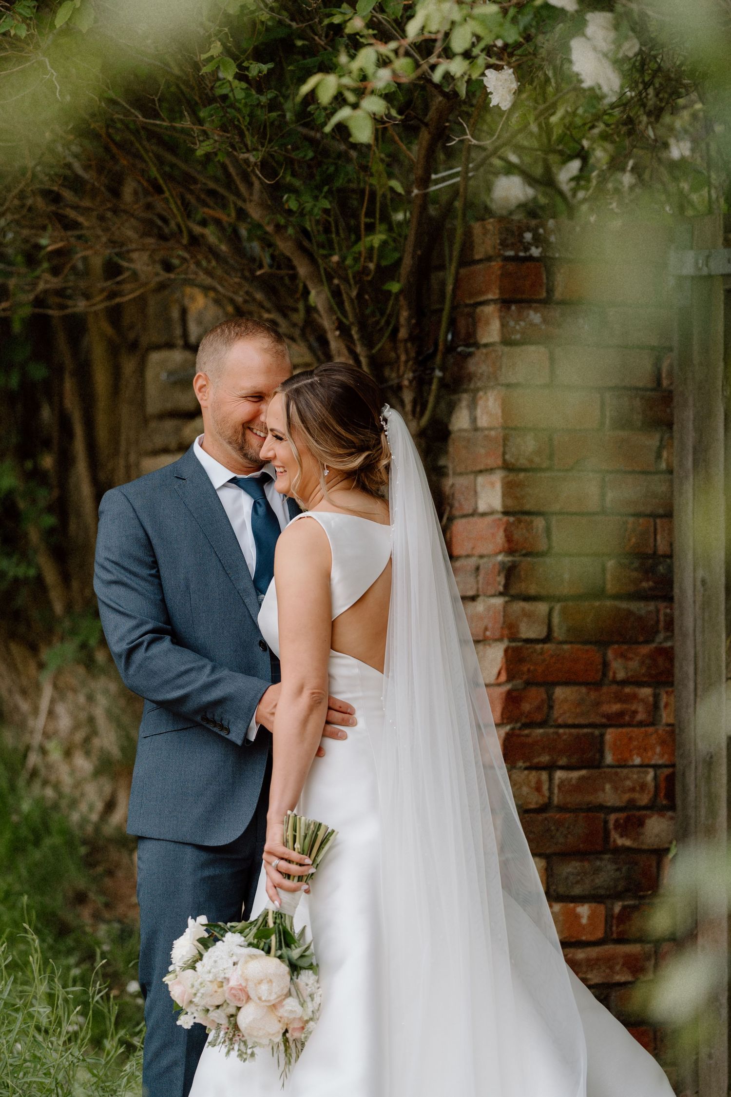 A romantic wedding moment captured outdoors with a bride in white gown holding flowers against a rustic brick wall backdrop at Dodford Manor