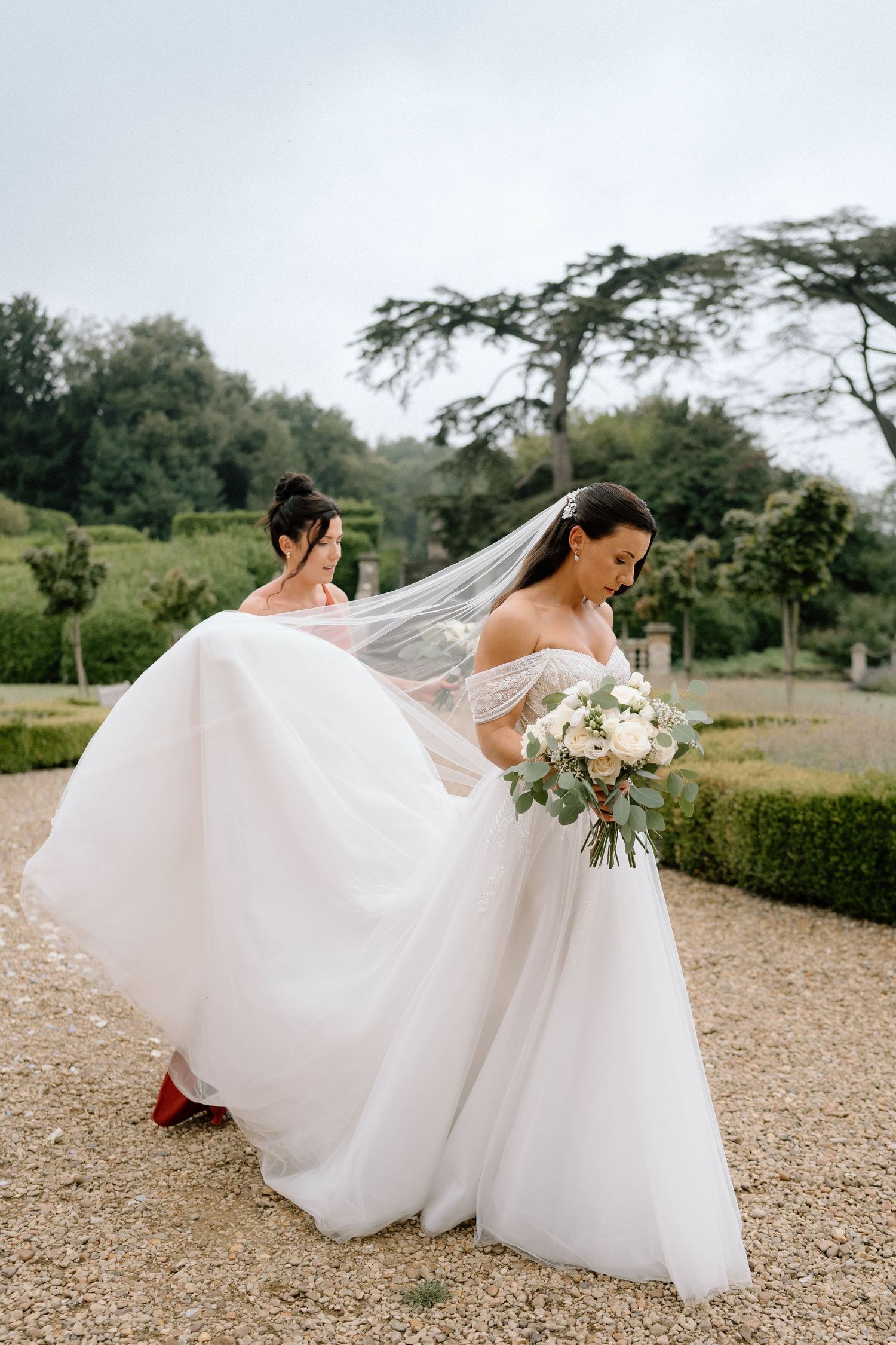 A bride in a flowing white gown holds a bouquet while her dress billows in the wind on a garden path at Harlaxton Manor.