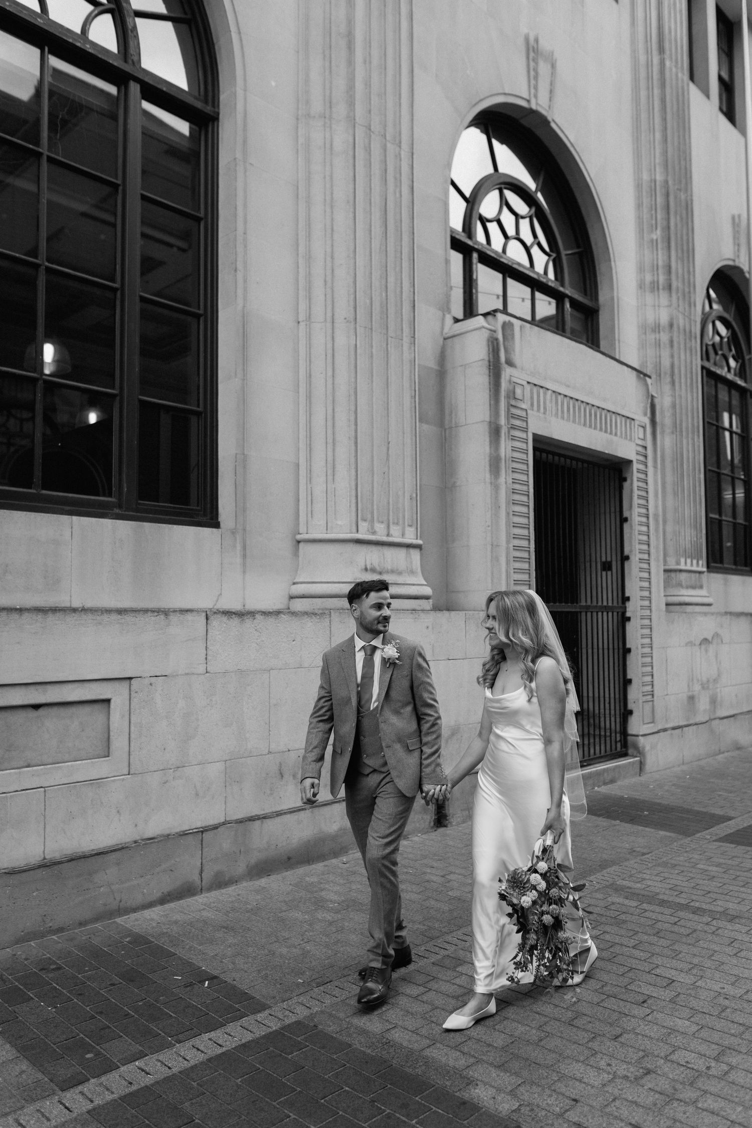 Black and white photo of a couple walking past classic architecture after their wedding ceremony in Coventry.