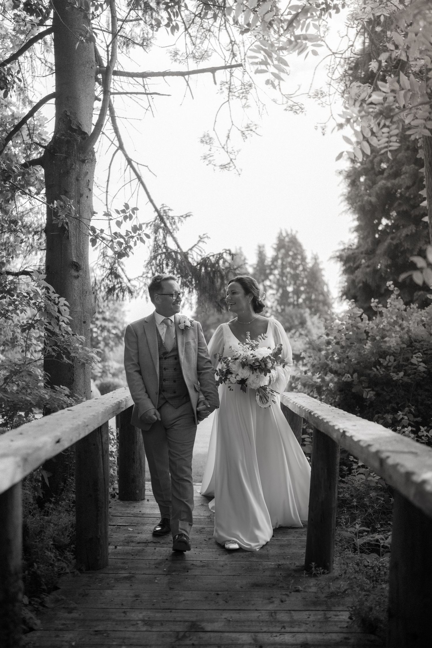 Black and white photo of couple walking across wooden bridge surrounded by trees at Nuthurst Grange.