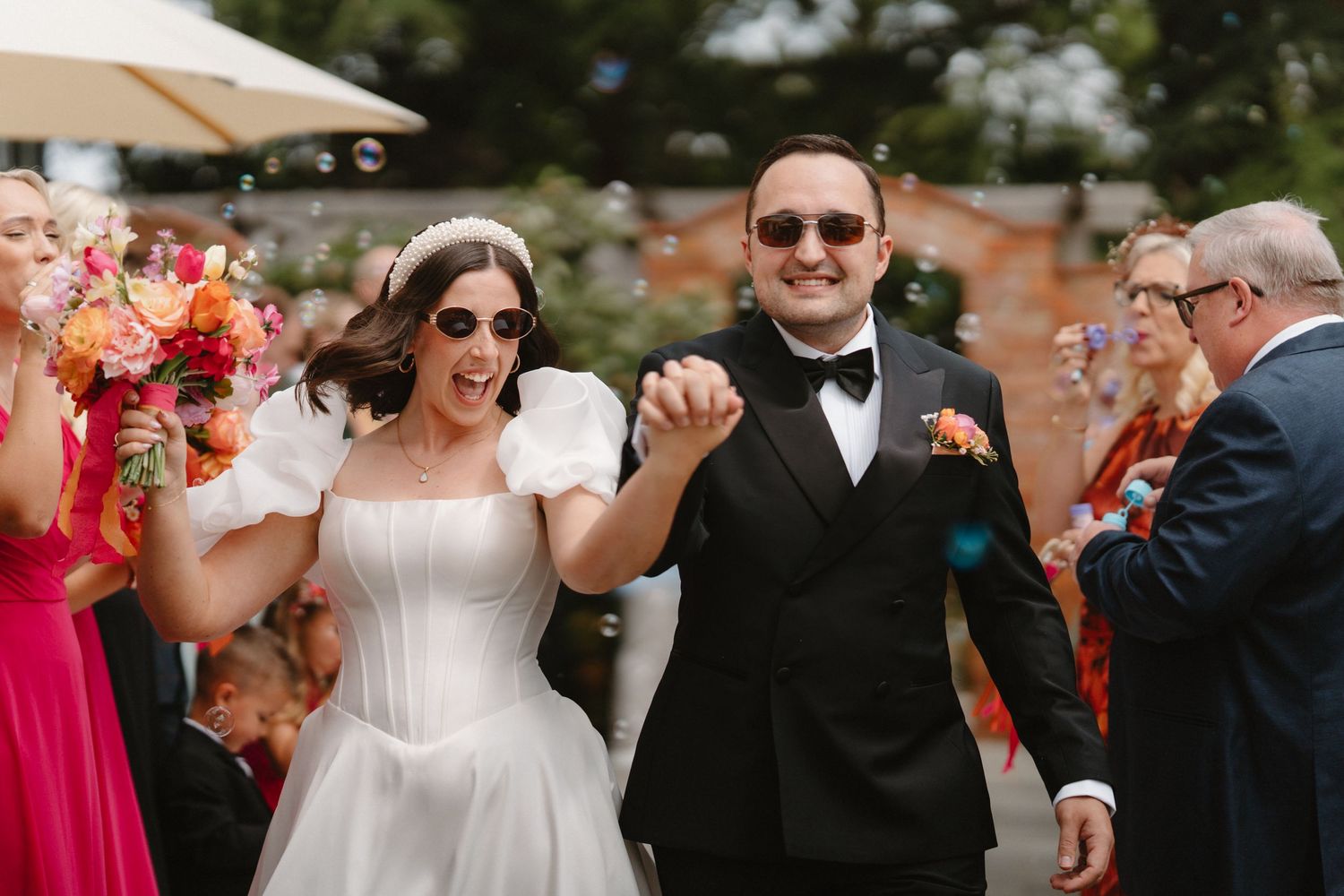 Joyful newlyweds in sunglasses walk through confetti shower, bride in white gown, groom in black tuxedo.