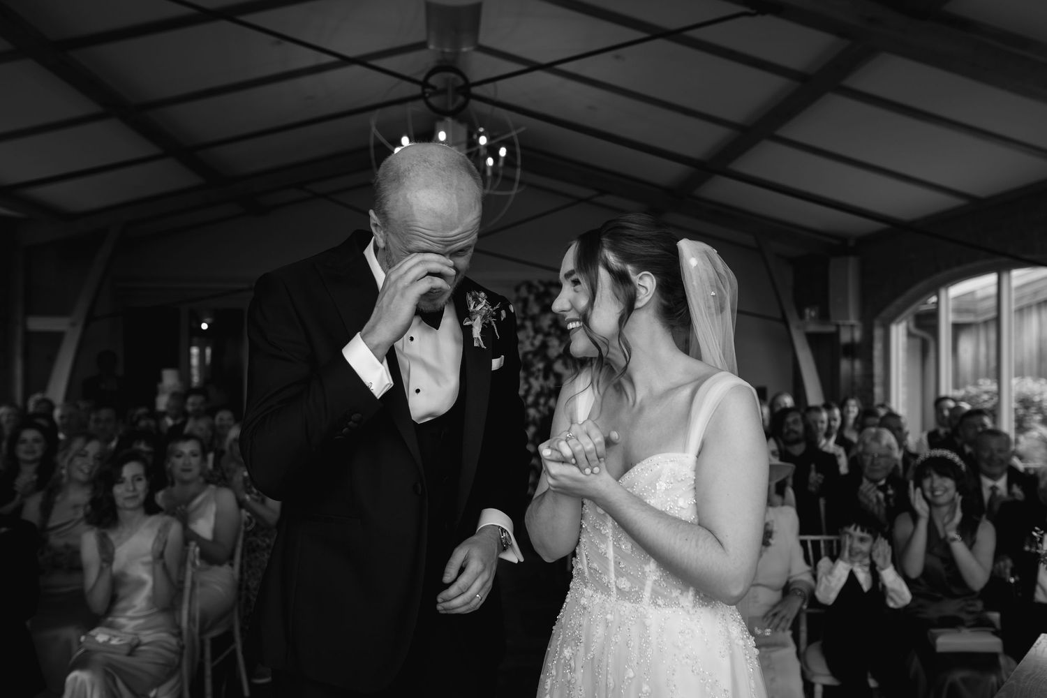 Emotional groom wiping tears while bride smiles during black and white wedding ceremony photo.