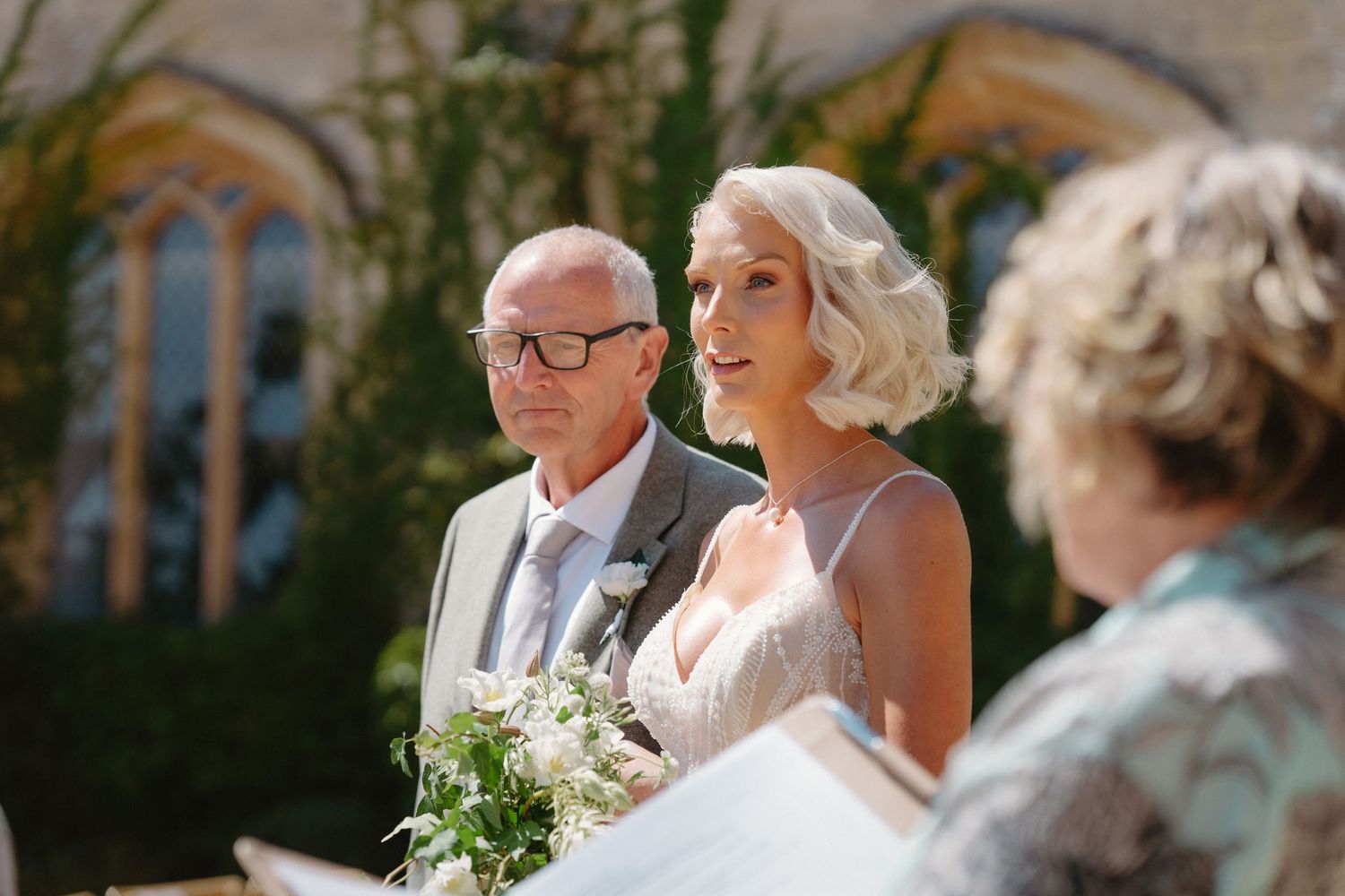 Bride holding bouquet stands with bald groom in gray suit during outdoor wedding ceremony at stone church.