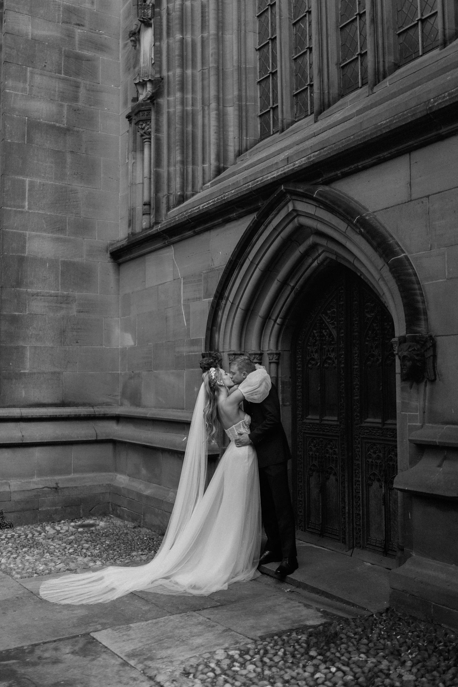 Romantic black and white photo of newlyweds embracing by gothic church doorway.