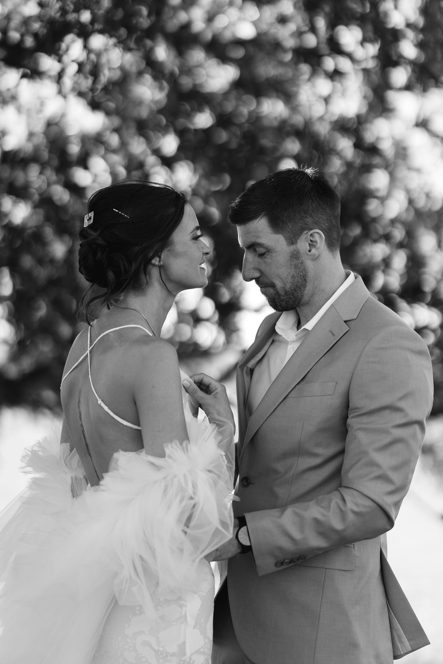 Black and white photo of bride and groom sharing an intimate moment outdoors, with bokeh trees in background.